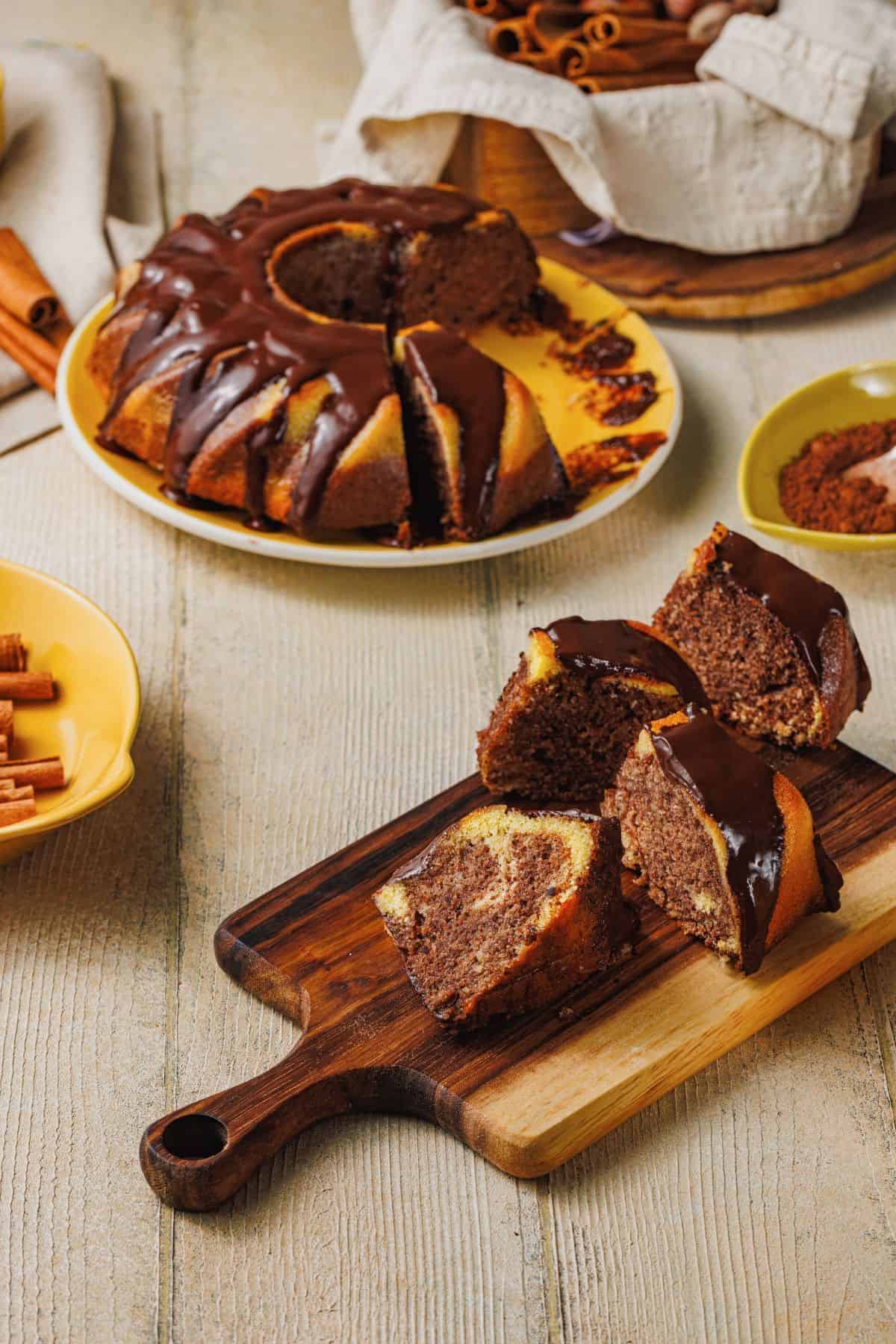 Slices of Cinnamon Marble Bundt Cake on a wooden board with the whole cake in a serving dish, surrounded by ingredients.