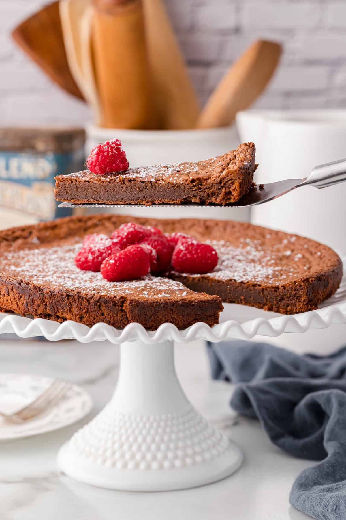 Flourless chocolate cake on a cake stand being sliced, topped with fresh raspberries.