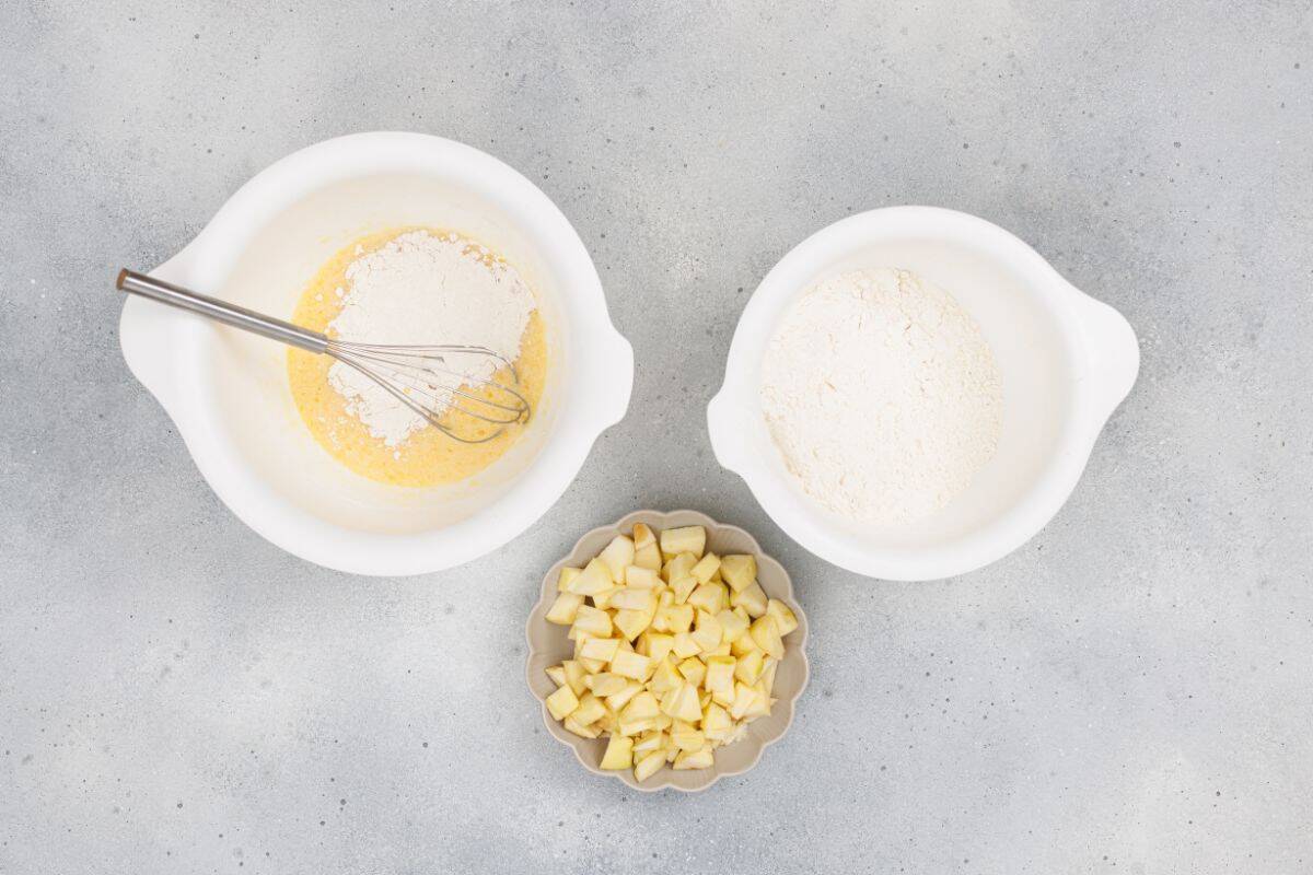 Two bowls containing dry and wet ingredients separately with diced apples placed nearby in a small bowl.