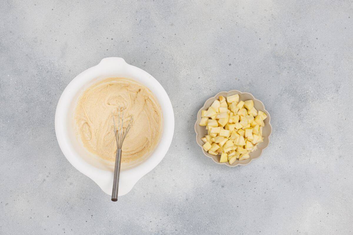 Mixed ingredients in a large bowl with a whisk resting on it and diced apples in a small bowl nearby.