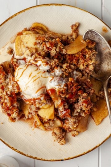 Apple crisp served on a plate topped with a scoop of vanilla ice cream, with a spoon on the side.