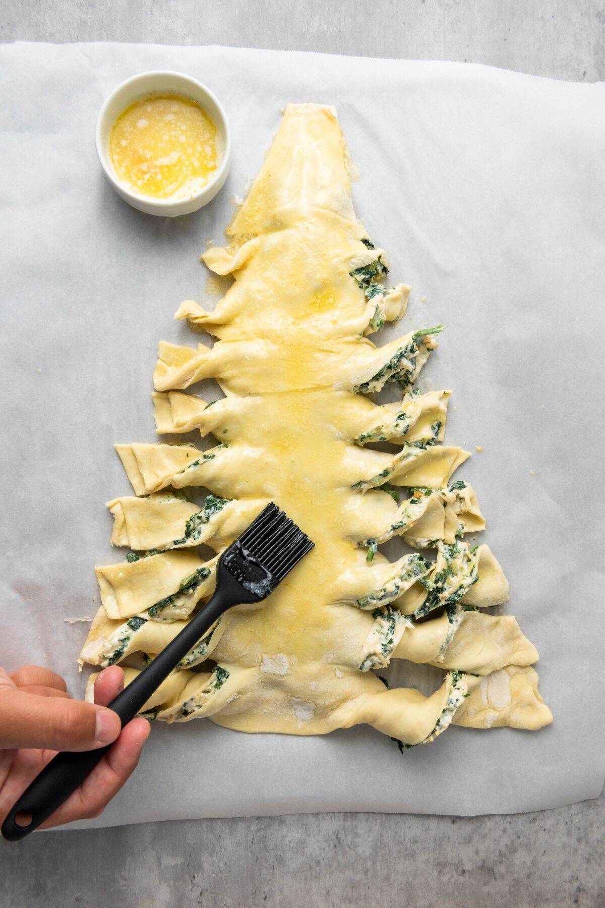 A hand brushing egg wash over the spinach dip pastry tree before baking.