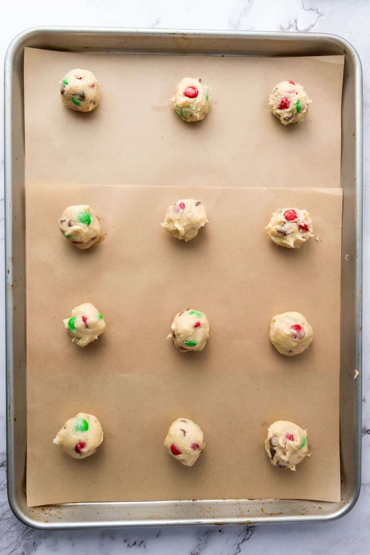 Scooped portions of Christmas M&M cookie dough lined up on a parchment paper-covered baking sheet, ready to bake.