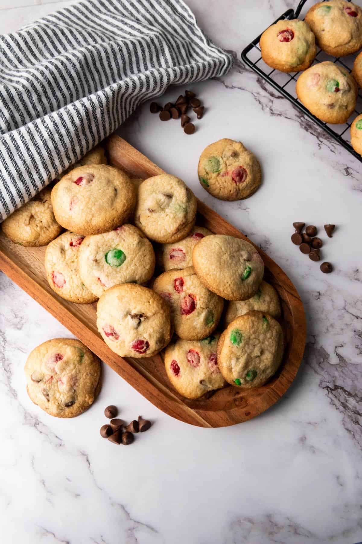 A batch of Christmas M&M cookies, some stacked neatly on a wooden serving board and others scattered on the surface around it.
