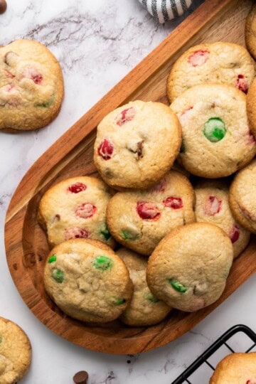 A batch of Christmas M&M cookies, some stacked neatly on a wooden serving board and others scattered on the surface around it.