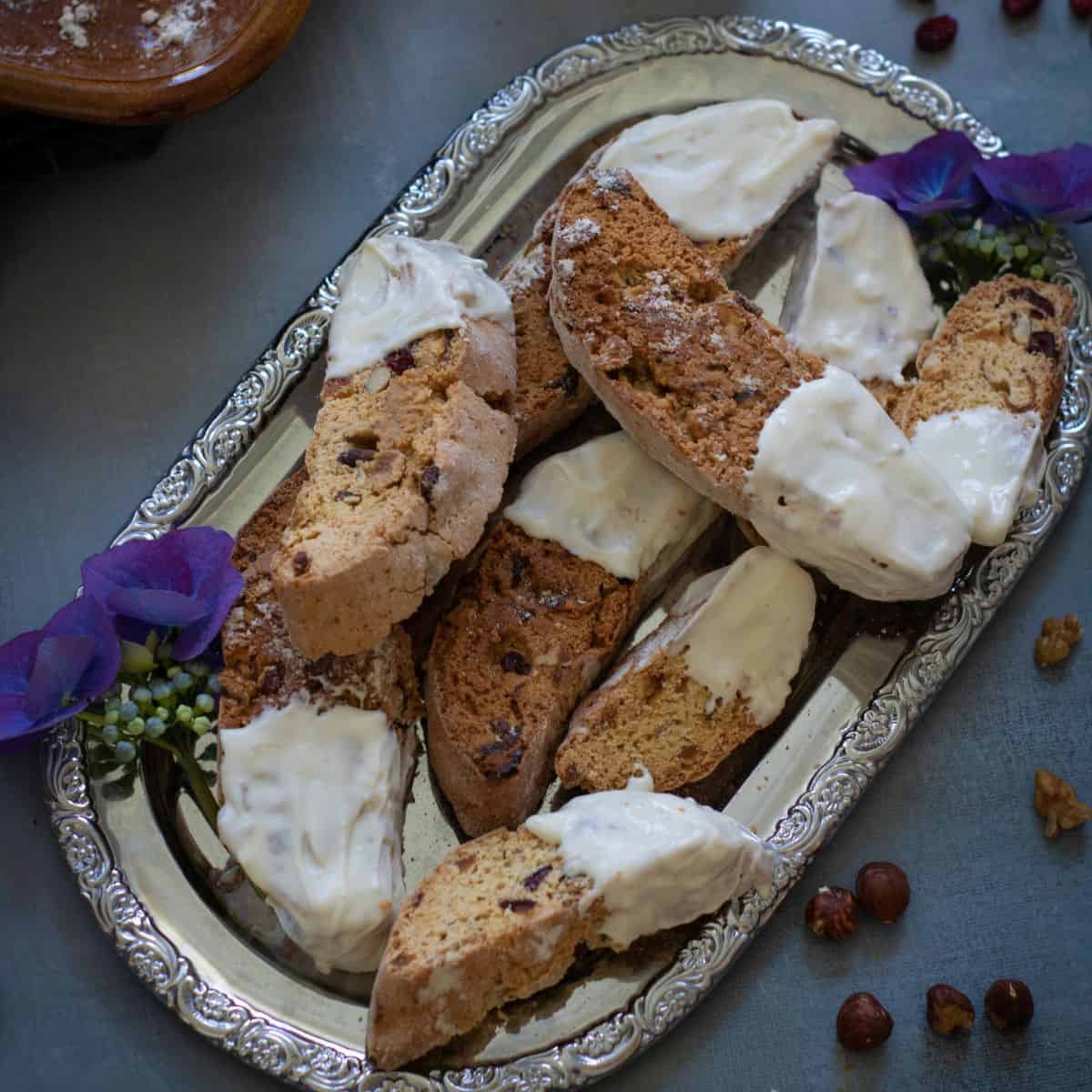 Finished cranberry apricot walnut biscotti dipped in white chocolate, arranged on a decorative tray, garnished with dried cranberries and orange zest.
