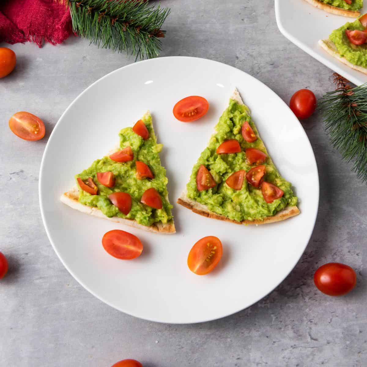 Festive avocado pita tree appetizers on a plate, surrounded by cherry tomatoes, with holiday-themed decorations in the background.