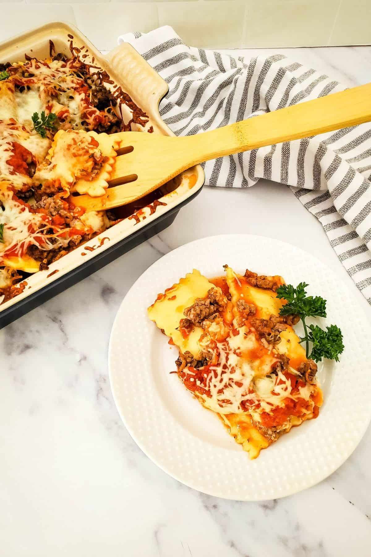 Cheesy ravioli lasagna bake served on a plate, with a larger portion in a serving dish beside it and a spatula resting on the dish.