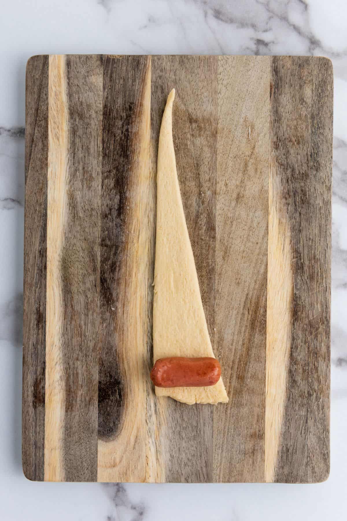 A single sausage placed in a small dough triangle on a wooden board.