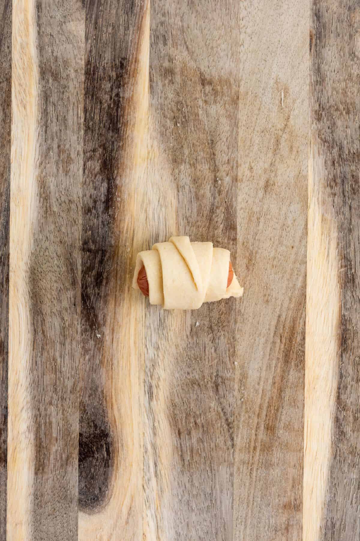 A single sausage being rolled inside a small dough triangle on a wooden board.