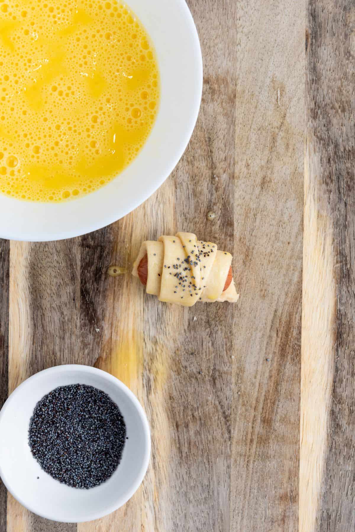 A single sausage being rolled inside a small dough triangle on a wooden board with egg wash bowl on the side, topped with sesame seeds.