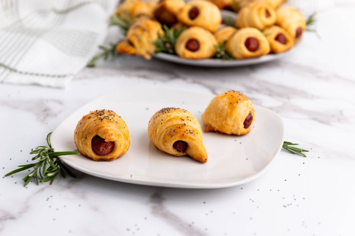 Mini piggies in a blanket served on a plate with many more blurred in the background.