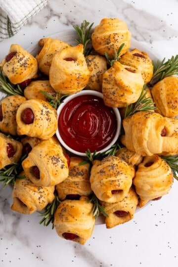 Mini piggies in a blanket arranged on a plate around a small bowl of dipping sauce, garnished with herbs.