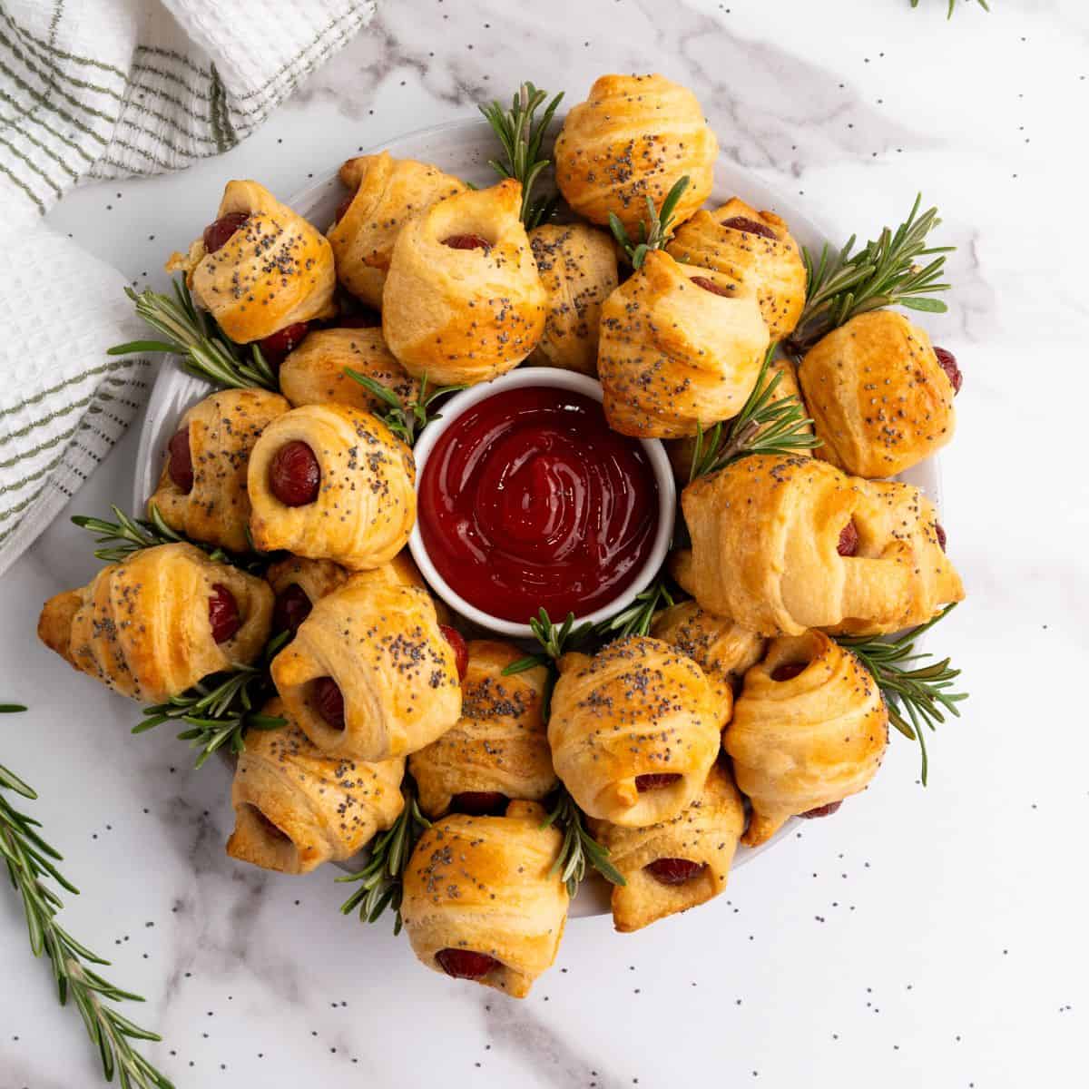 Mini piggies in a blanket arranged on a plate around a small bowl of dipping sauce, garnished with herbs.