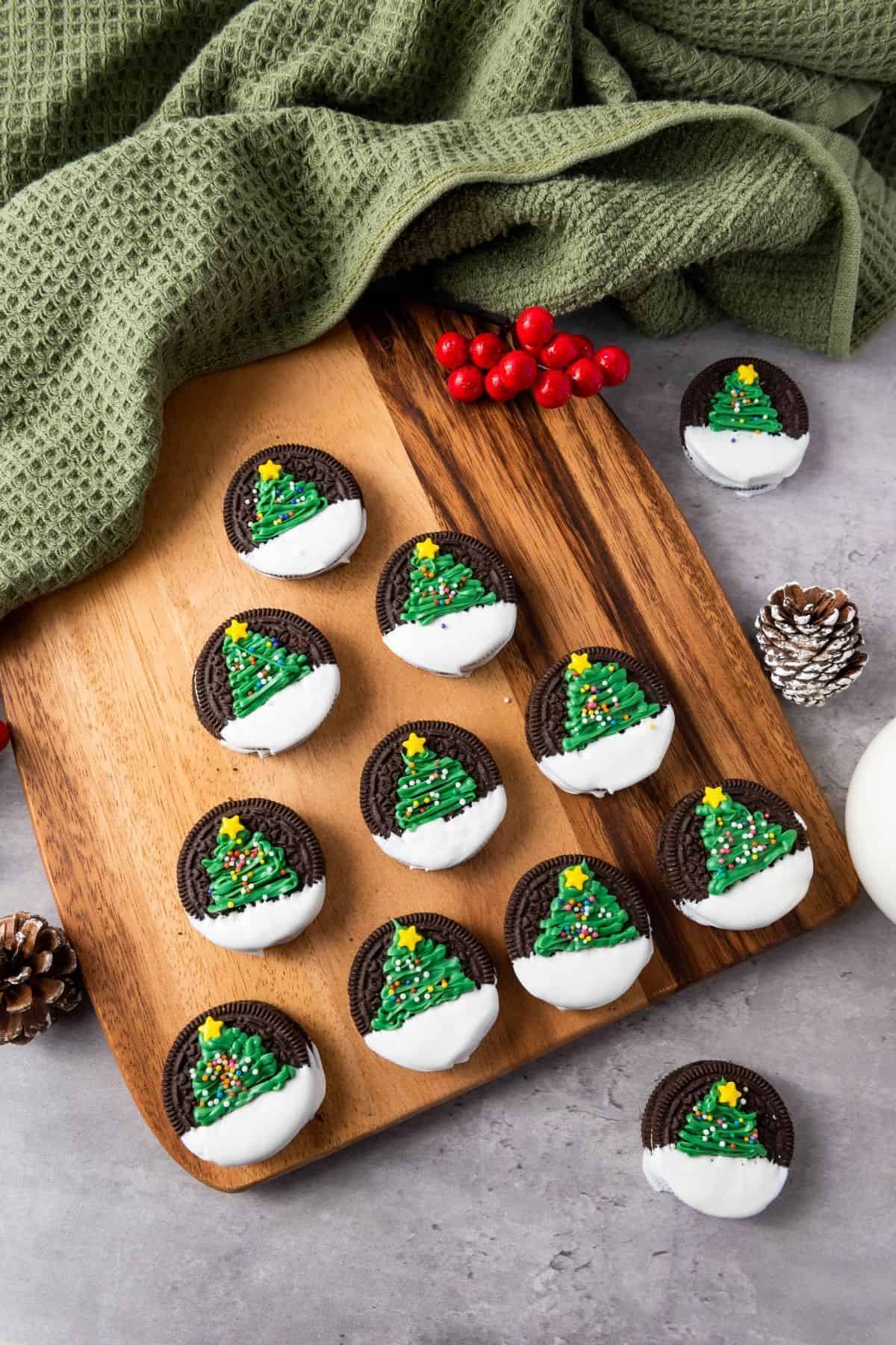 Cookies topped with Christmas tree decorations, neatly arranged on a wooden board and surrounded by festive Christmas ornaments.