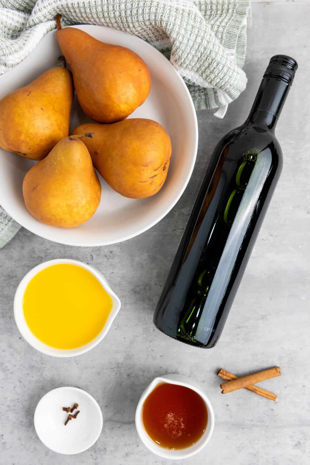 Red wine, orange juice, honey, cinnamon sticks, cloves, and four Bosc pears arranged neatly on a kitchen surface, ready for poaching.
