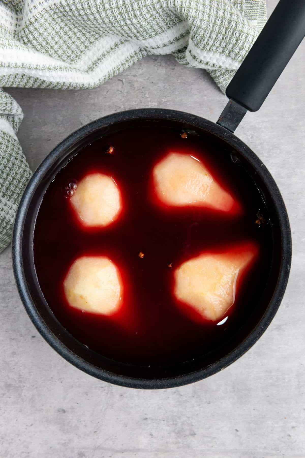 A pear being soaked in a red wine and spice mixture in a saucepan, ready for poaching.