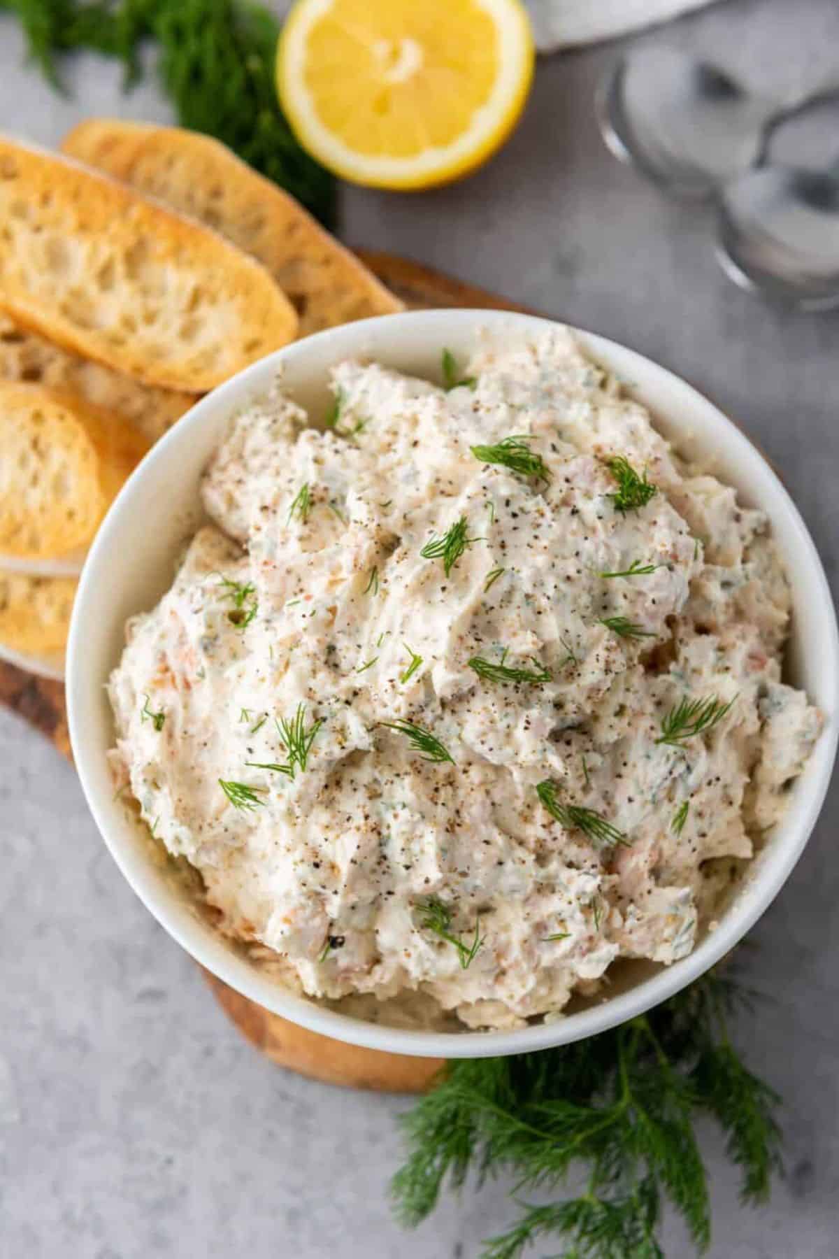 A bowl of smoked salmon dip garnished with a slice of lemon, served with pieces of garlic bread on the side.