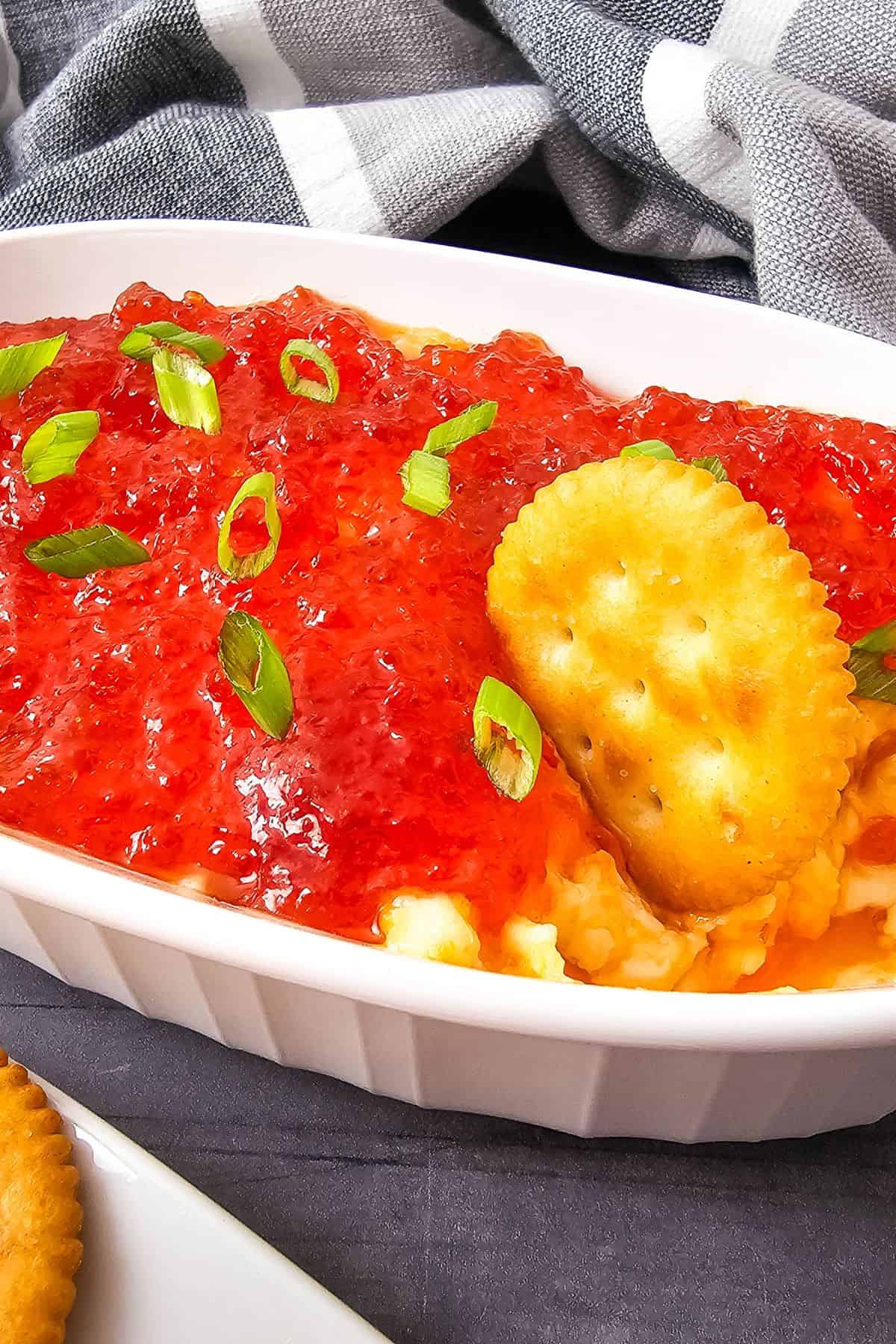A serving dish filled with pepper jelly cheese dip topped with glossy red jelly, with a biscuit resting on the edge ready for dipping.
