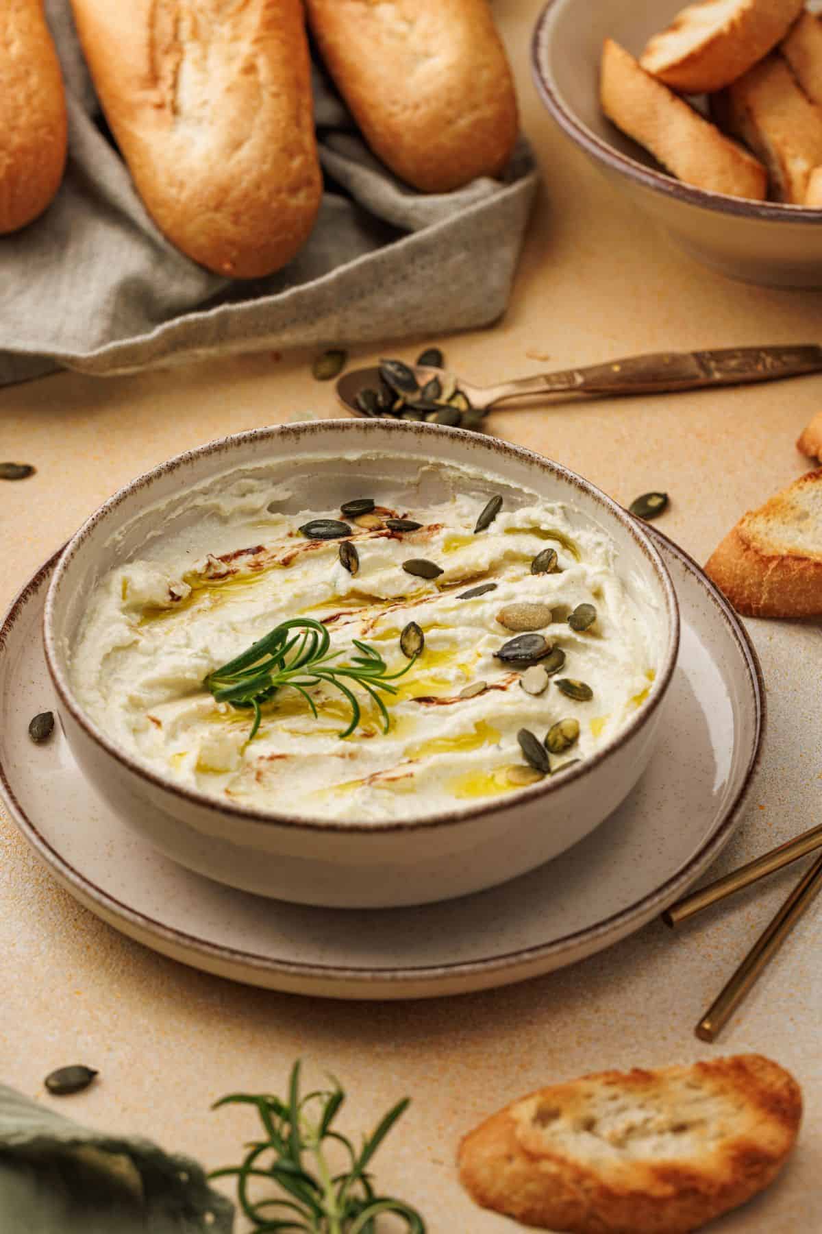A bowl of creamy baked goat cheese dip garnished with fresh herbs and crushed nuts, ready to be served with crackers or bread on the side.
