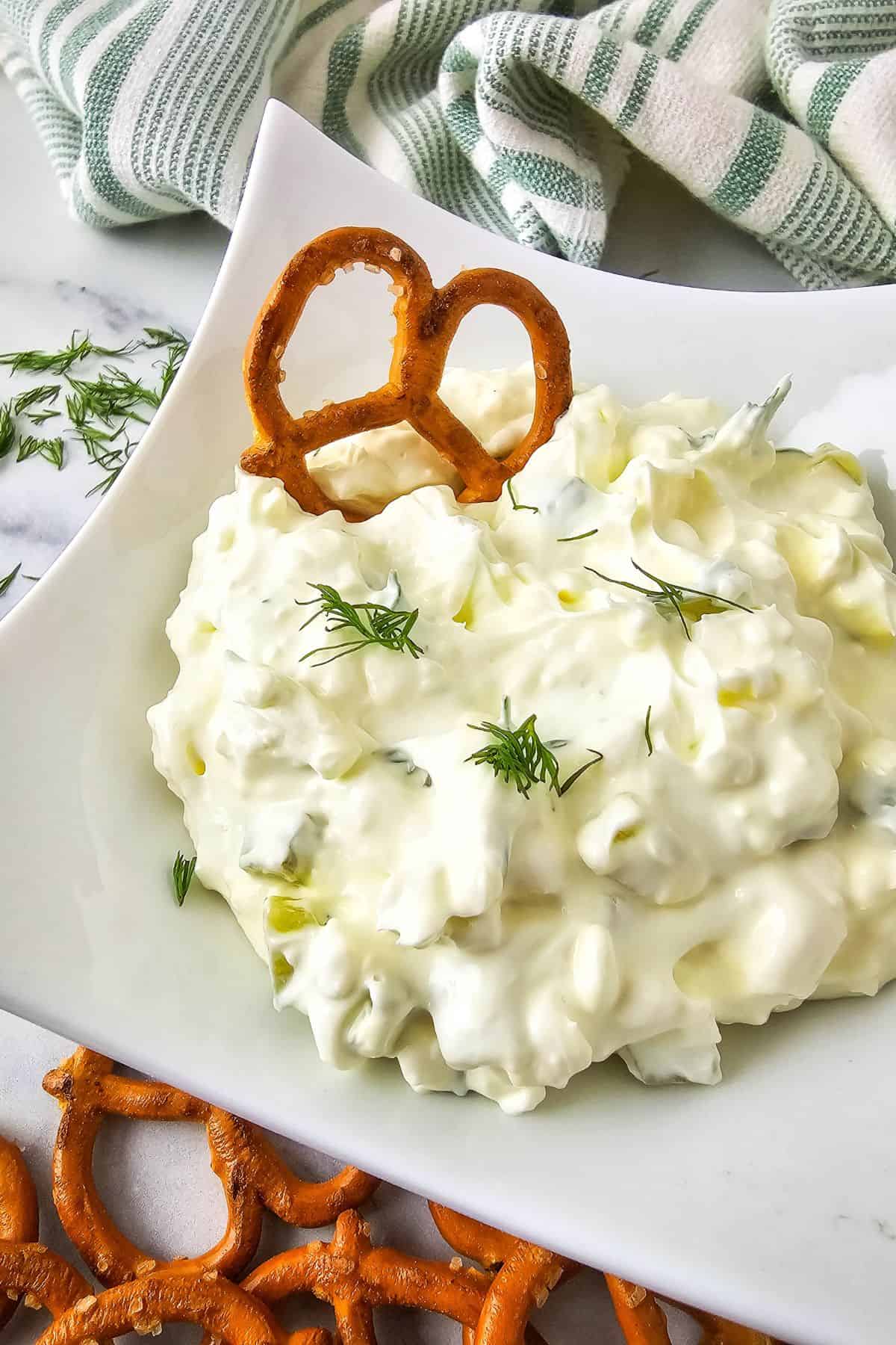 A serving plate filled with creamy dill pickle dip topped with chopped pickles, with pretzels resting on the side for dipping.