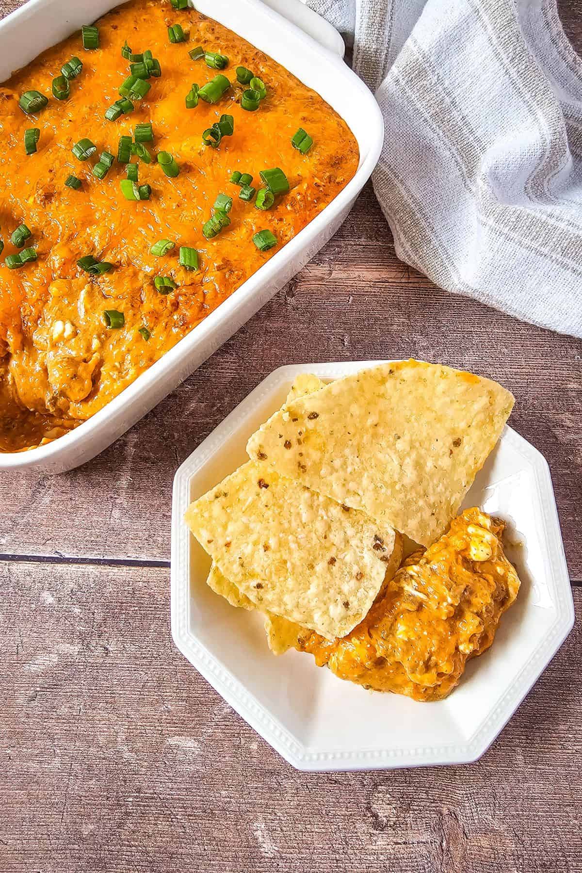 Loaded chili cheese dip in a serving dish with melted cheese on top, served alongside tortilla chips and a small plate holding a portion of the dip.