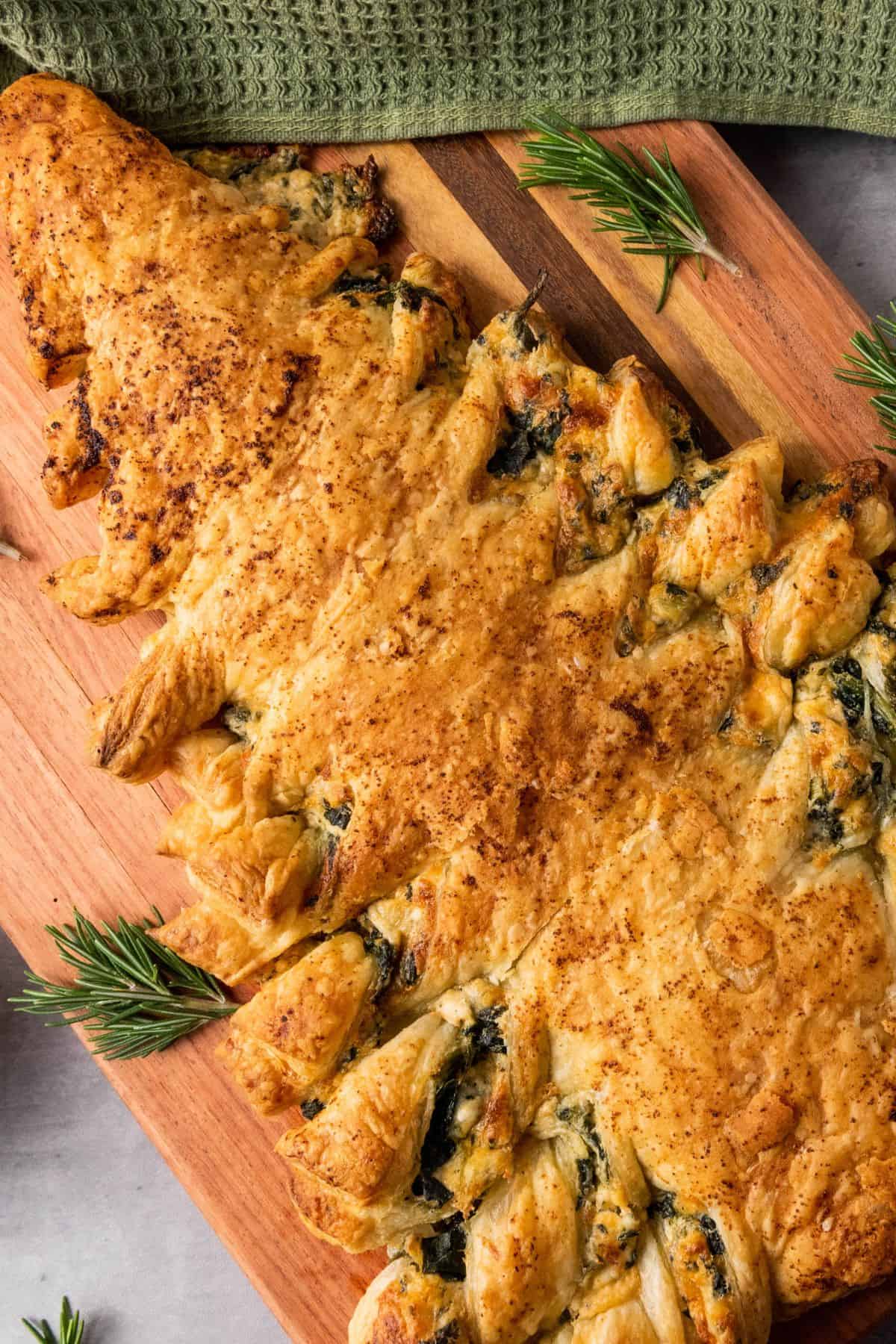 A wooden board displaying a cheesy spinach dip Christmas tree pastry with a table napkin on the side.