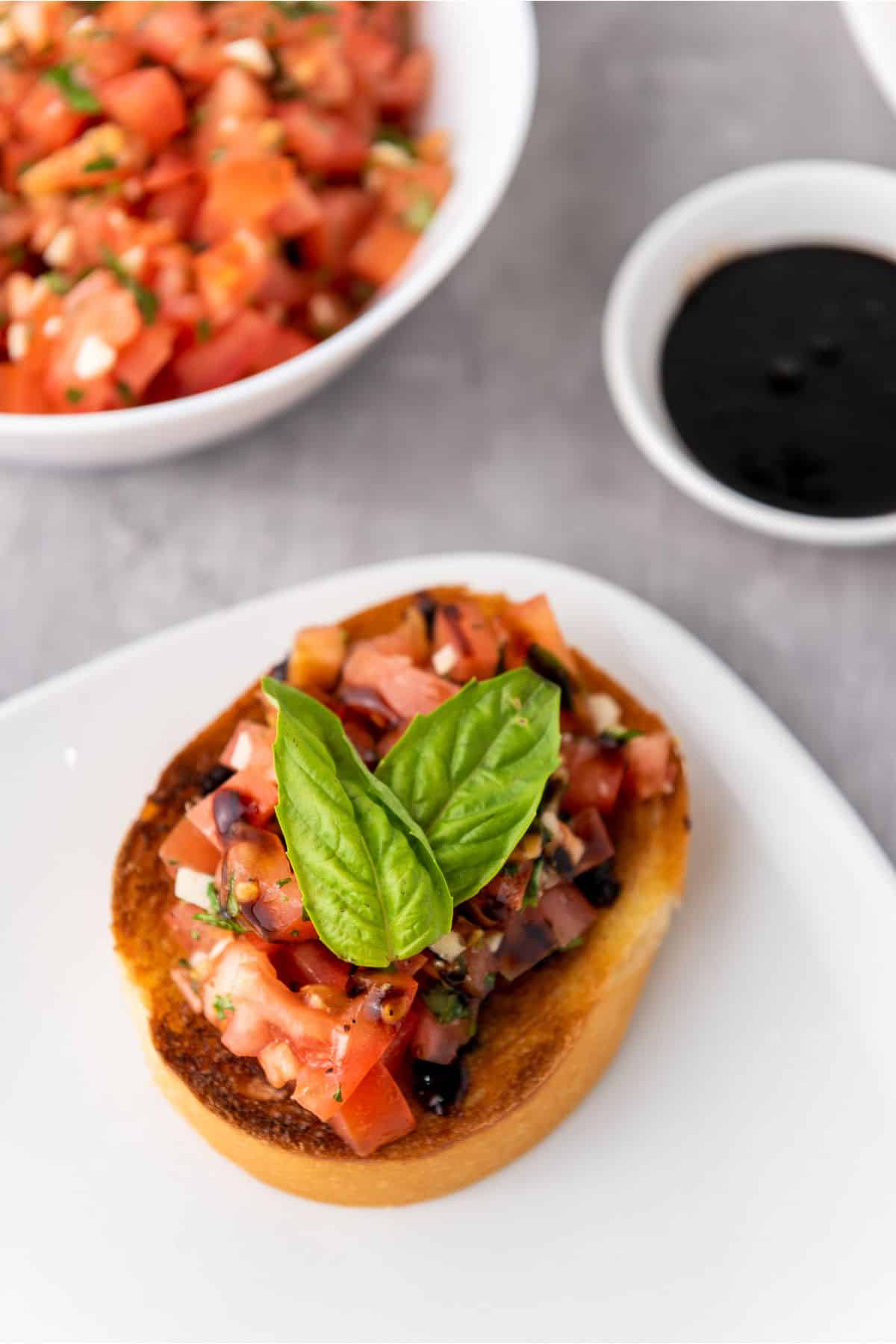 A single classic Mediterranean bruschetta served on a plate, topped with diced tomatoes and fresh basil, with a small ramekin of balsamic glaze on the side.