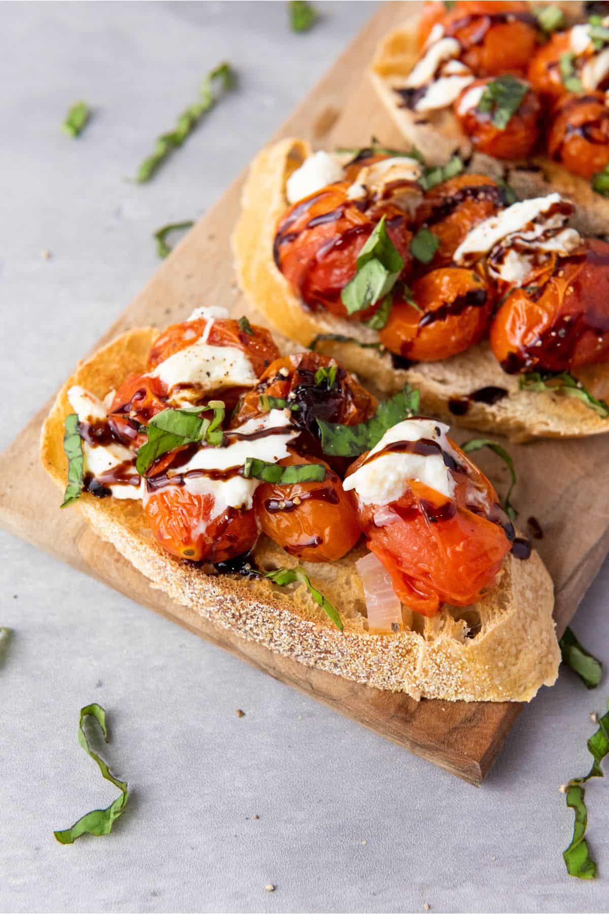 Roasted cherry tomato and burrata bruschetta arranged on a wooden board, garnished with fresh basil and a drizzle of balsamic glaze.
