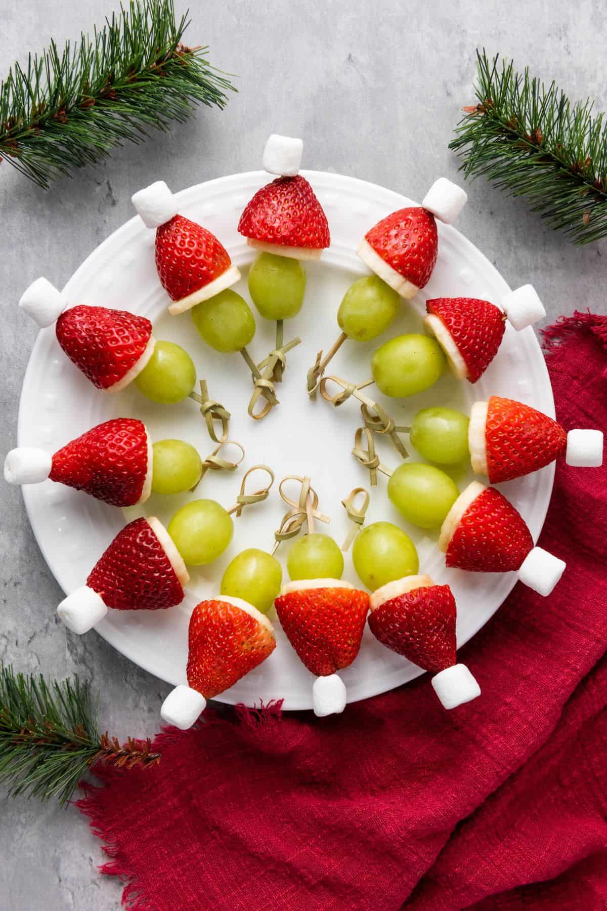Festive fruit kabobs arranged on a plate, featuring green grapes, strawberries, bananas, and mini marshmallows threaded onto skewers.
