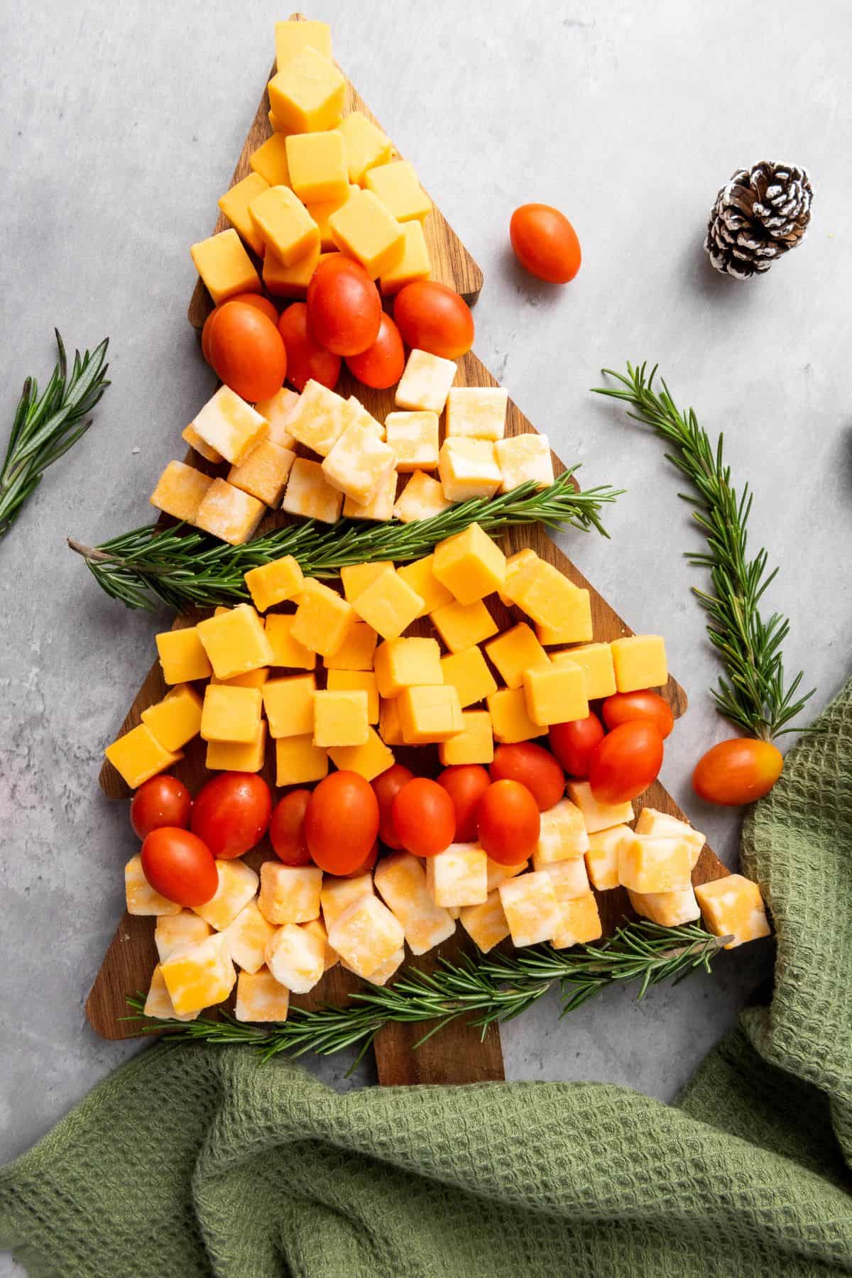 A Christmas tree-shaped cheese board arranged on a wooden board, decorated with assorted cheeses and garnished with fruits and nuts, with a table napkin on the side.
