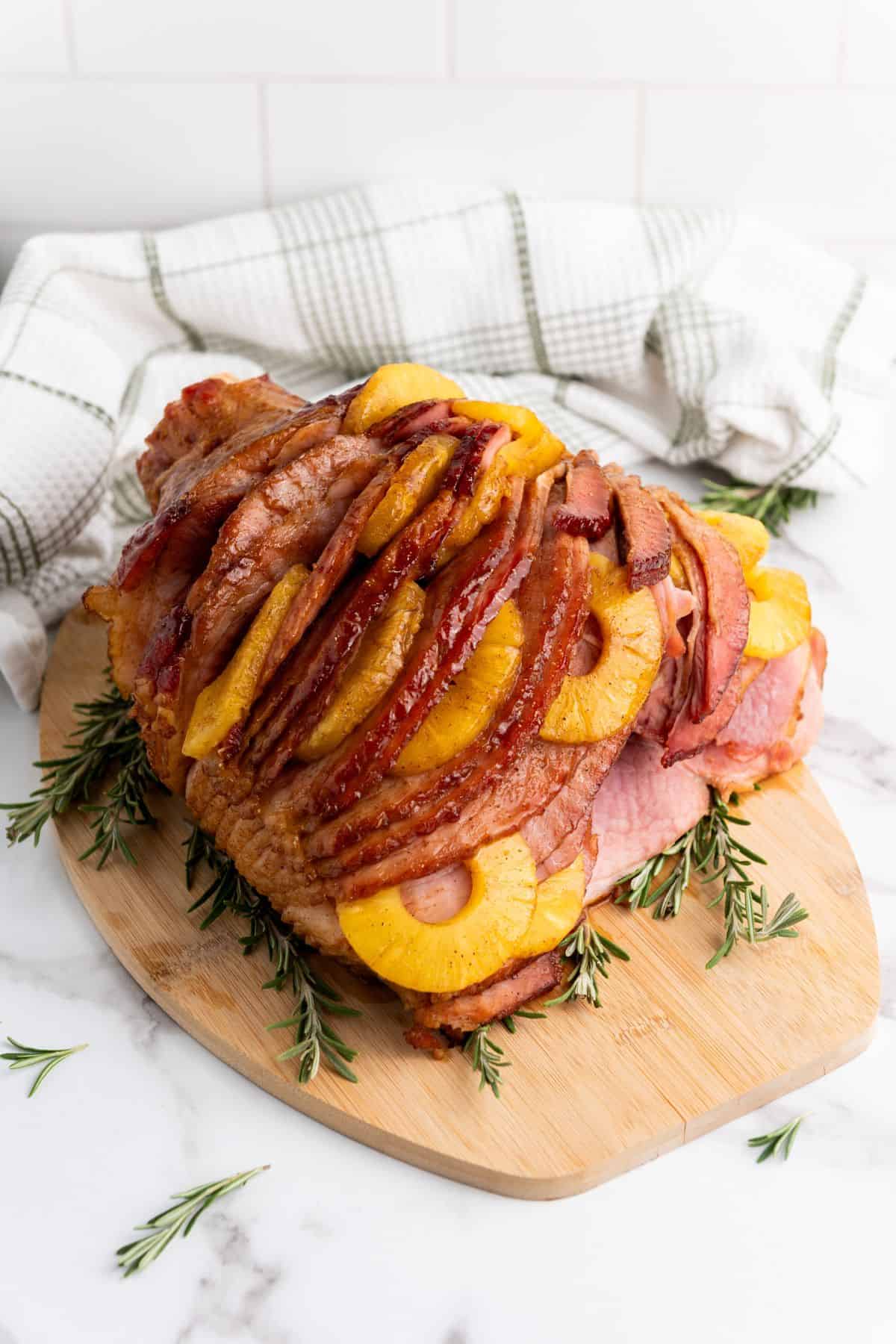 A whole brown sugar glazed ham topped with pineapple slices and garnished with fresh herbs, served on a wooden board.