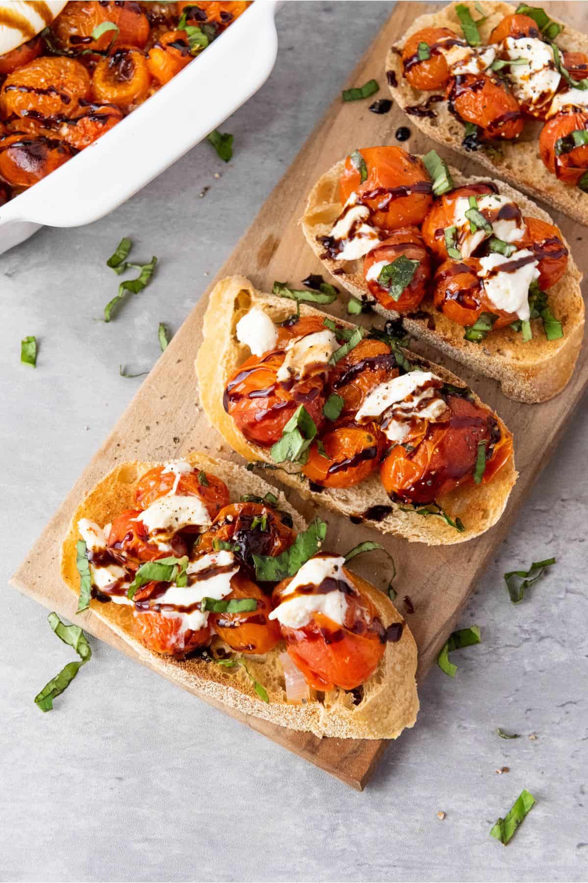 Roasted cherry tomato and burrata bruschetta arranged on a wooden board, garnished with fresh basil and a drizzle of balsamic glaze.