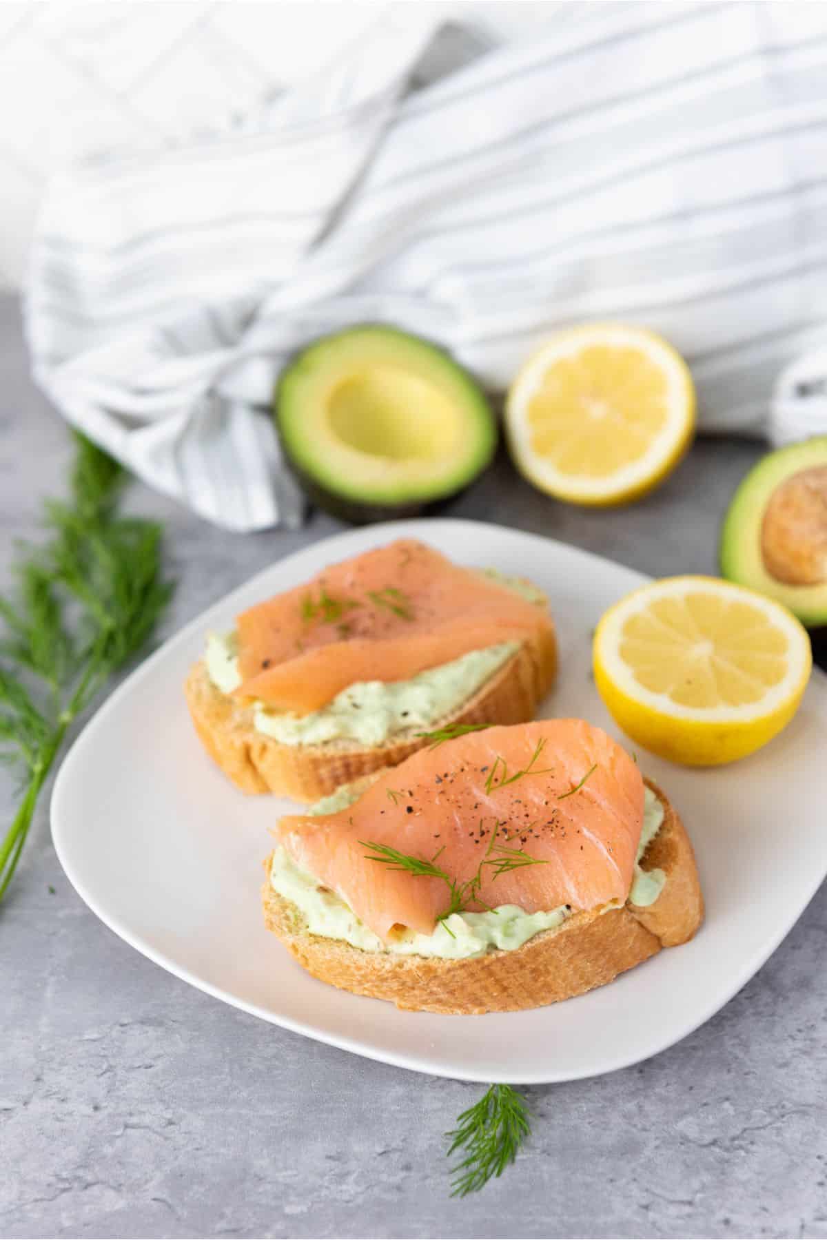 Smoked salmon and avocado bruschetta served on a plate, with avocado and lemon slices in the background.