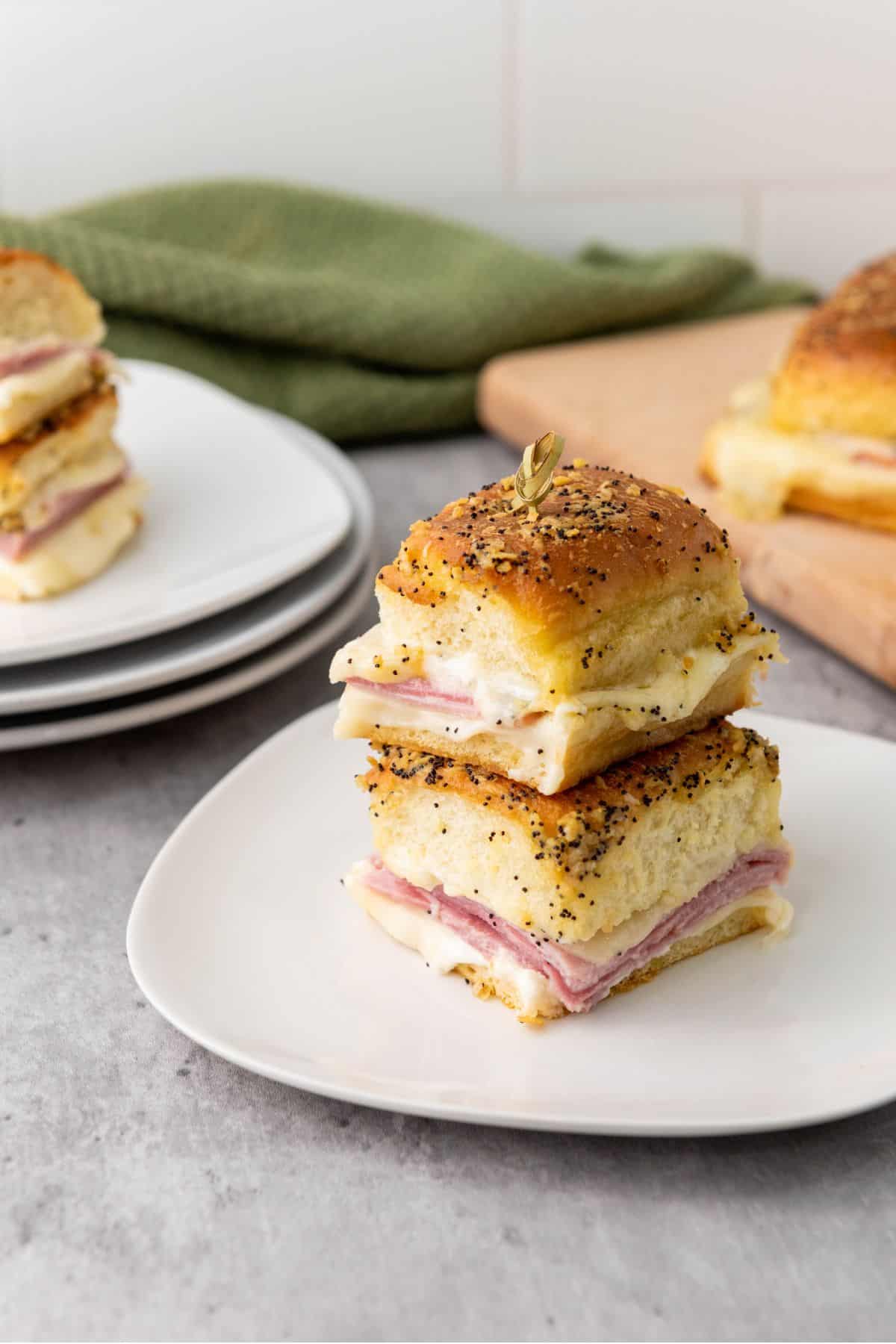 Stacked melty ham and cheese sliders on a plate, with additional sliders blurred in the background.