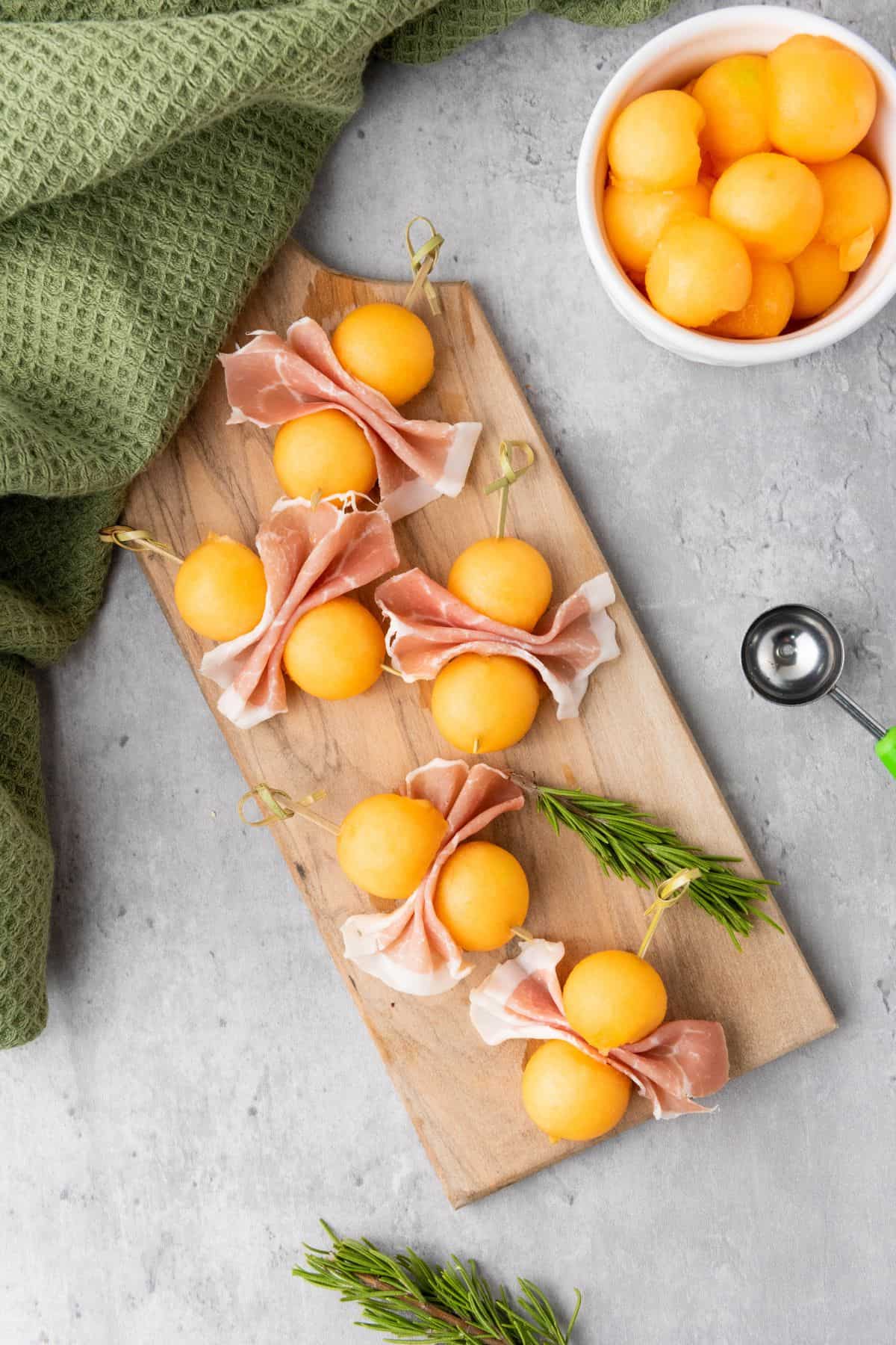 Cantaloupe prosciutto skewers neatly arranged on a rustic serving board with a table napkin on the side.