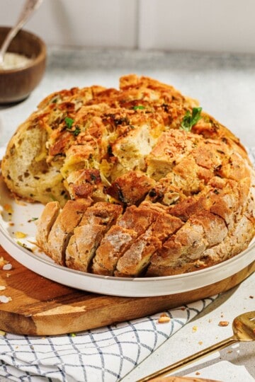 A golden, cheesy pull-apart garlic bread, partially sliced and garnished with fresh parsley, served on a plate placed on a wooden board, with melted cheese oozing between the cuts.
