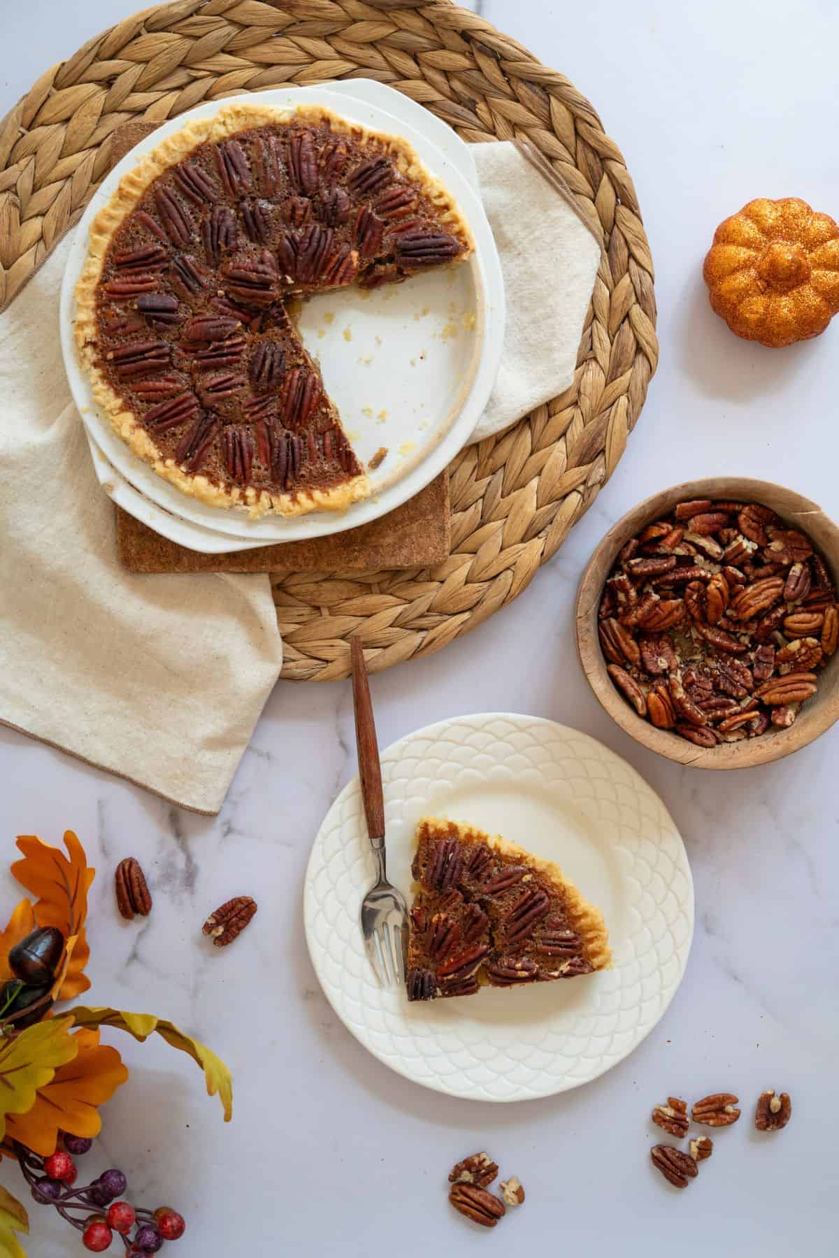 Top-down view of a pie in a dish with a slice served on a plate, accompanied by a fork and a small bowl of pecans.