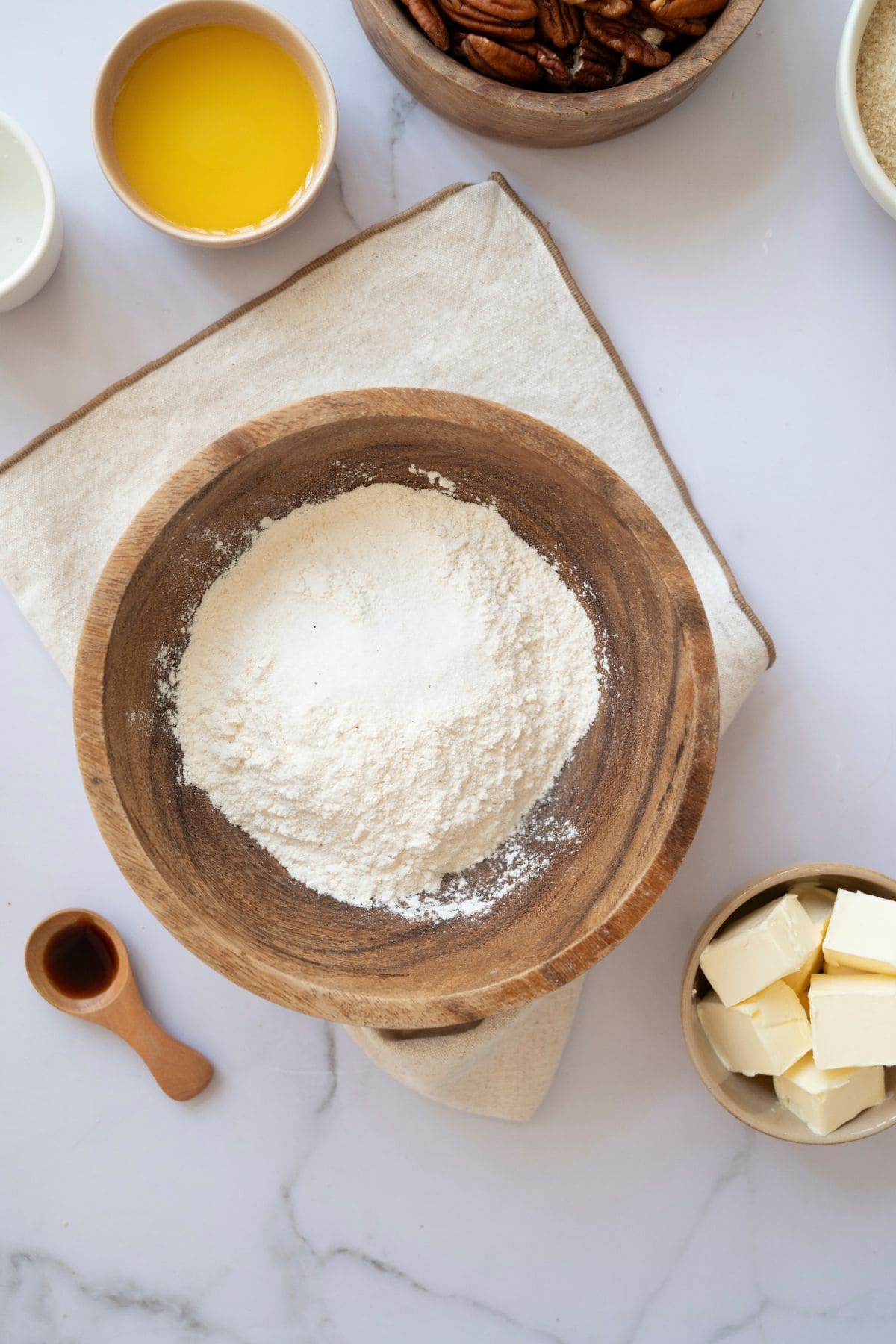 Flour in a large wooden bowl placed on a white tablecloth, surrounded by other baking ingredients.