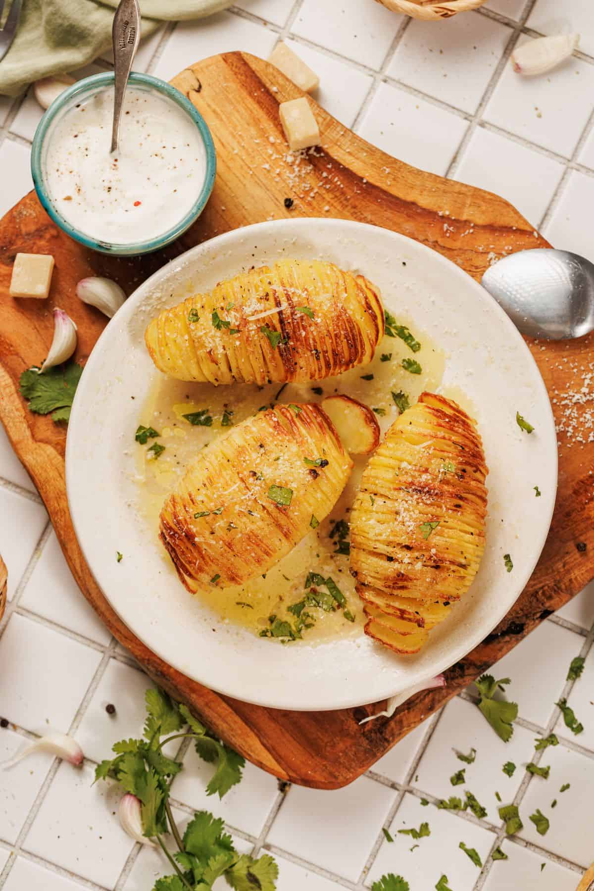 A plate with hasselback potato on a plate soaked in garlic-parmesan butter, topped with grated parmesan and fresh herbs, served with a creamy garlic dip.