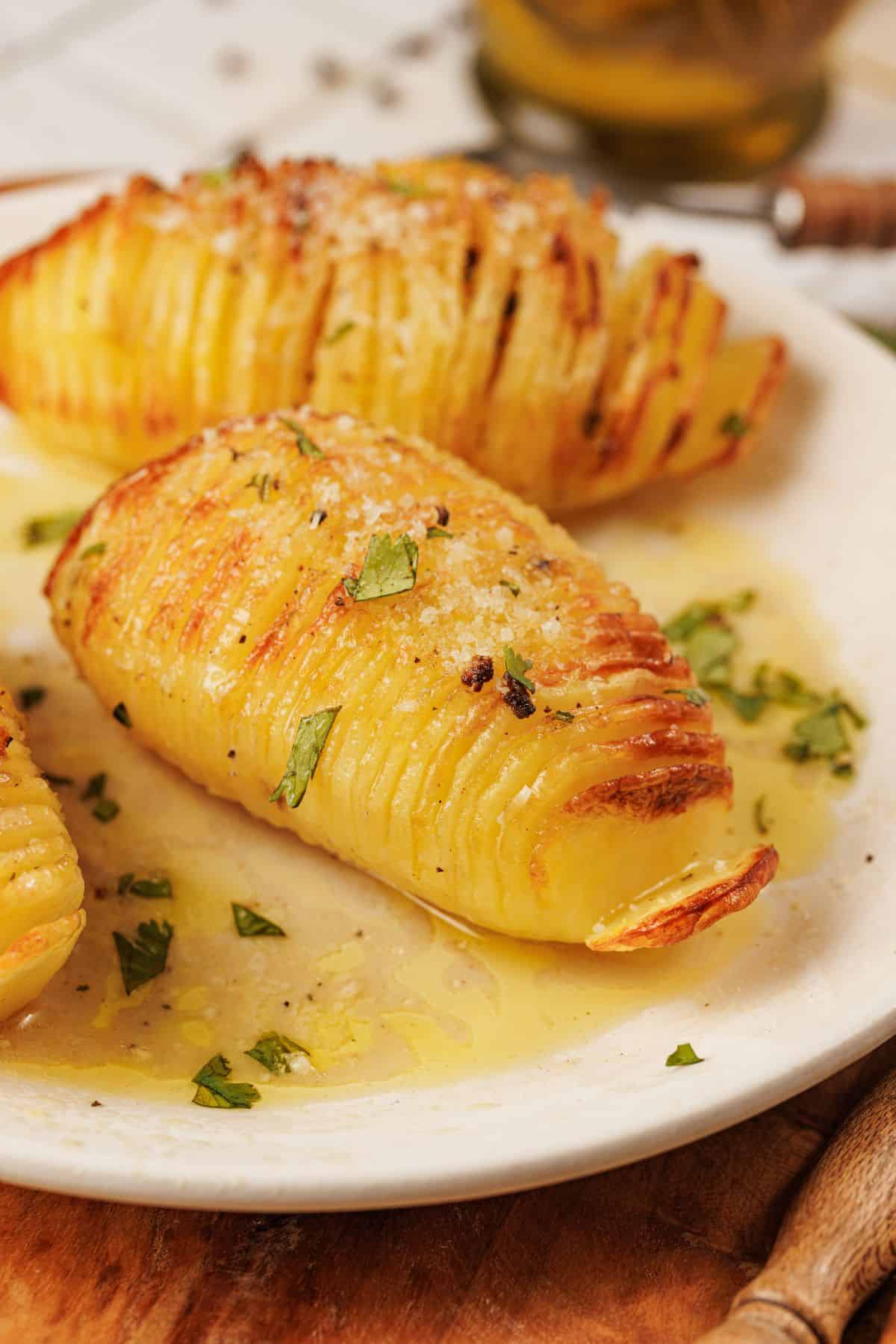 Close-up photo of hasselback potato on a plate soaked in garlic-parmesan butter, topped with grated parmesan and fresh herbs, served with a creamy garlic dip.