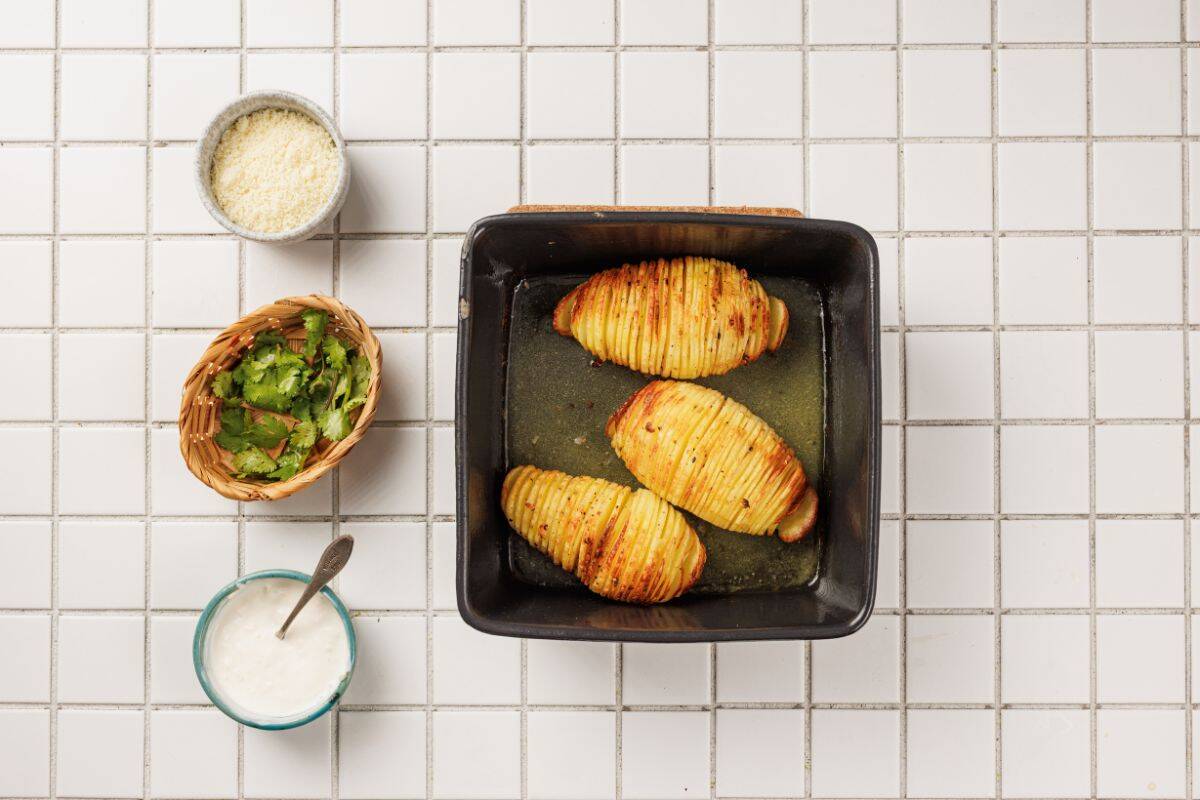 ooked Hasselback potatoes in a container surrounded by ingredients including garlic, butter, olive oil, Parmesan cheese, and fresh herbs.
