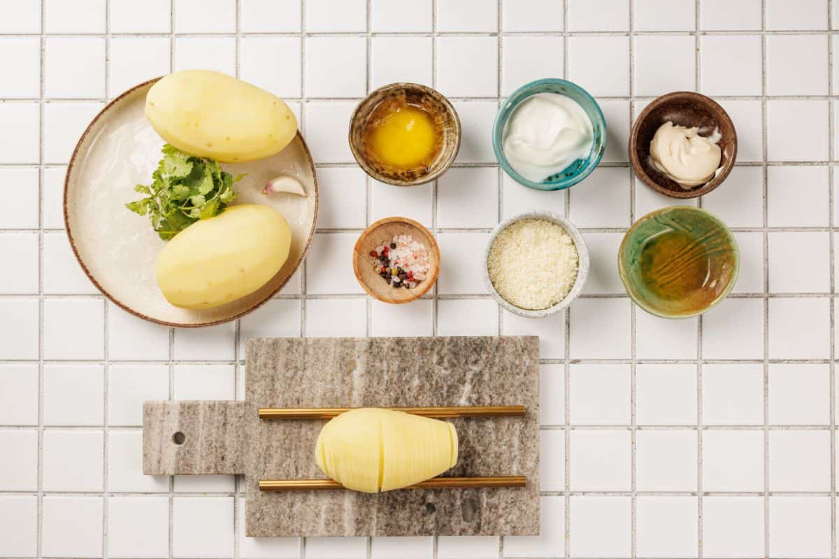 Potato placed on a cutting board surrounded by ingredients including garlic, butter, olive oil, Parmesan cheese, and fresh herbs.