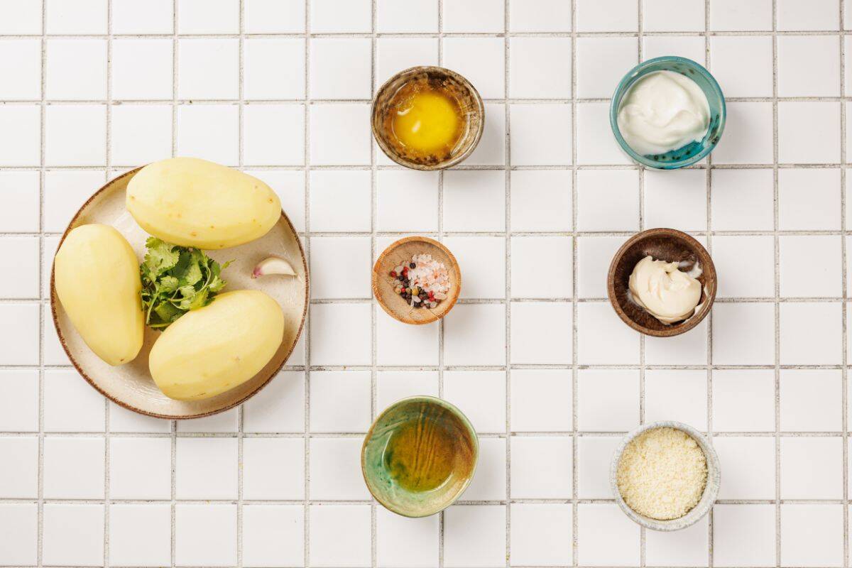Ingredients for Hasselback potatoes placed on a surface, including potatoes, garlic, butter, Parmesan cheese, olive oil, and fresh herbs.