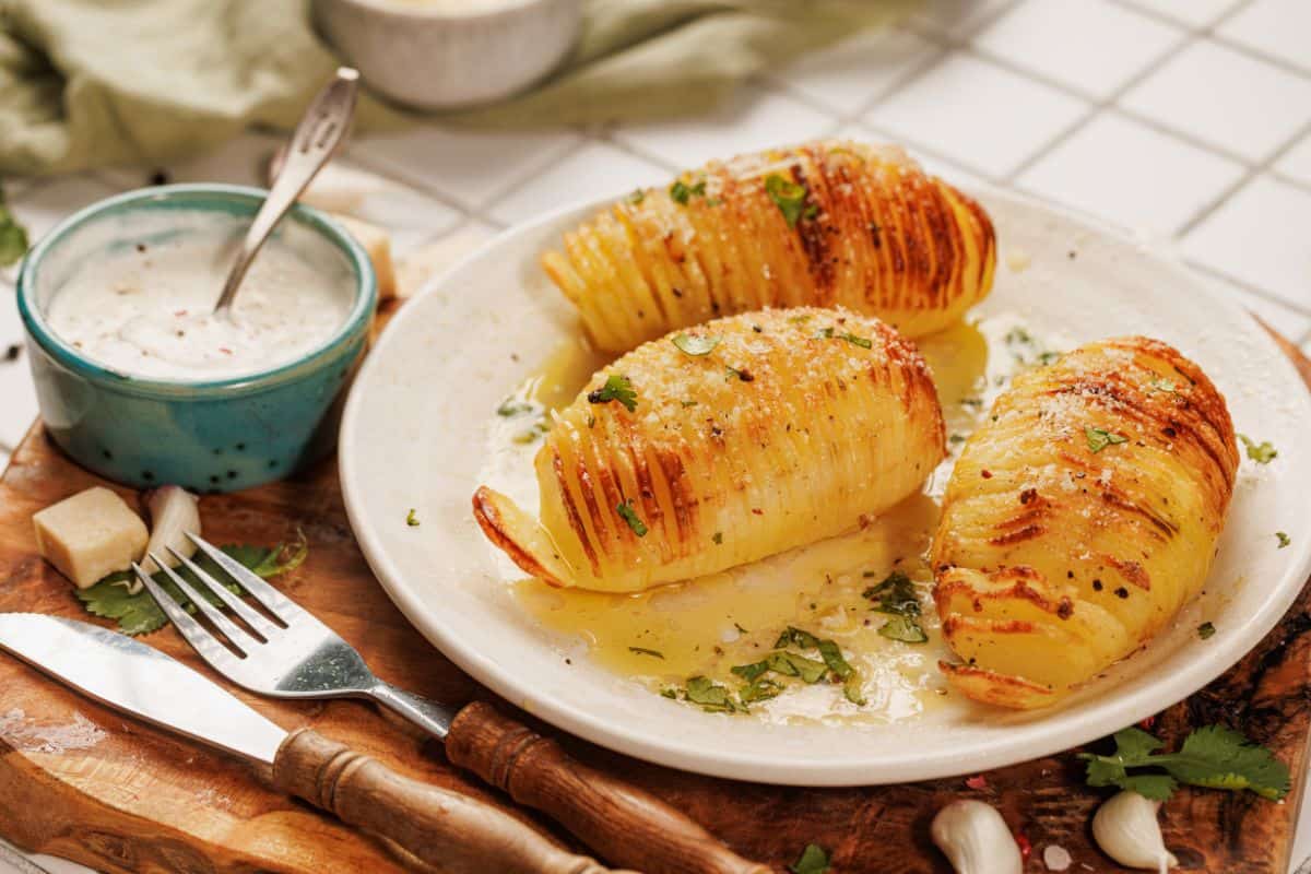 A plate with hasselback potato on a plate soaked in garlic-parmesan butter, topped with grated parmesan and fresh herbs, served with a creamy garlic dip.