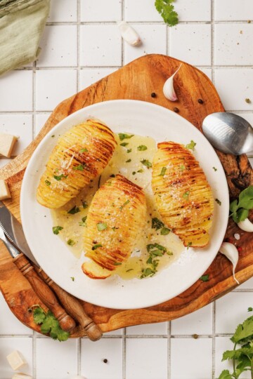 Hasselback potato served on a plate, garnished with grated Parmesan and fresh herbs, drizzled with garlic-parmesan butter, with utensils on the side.