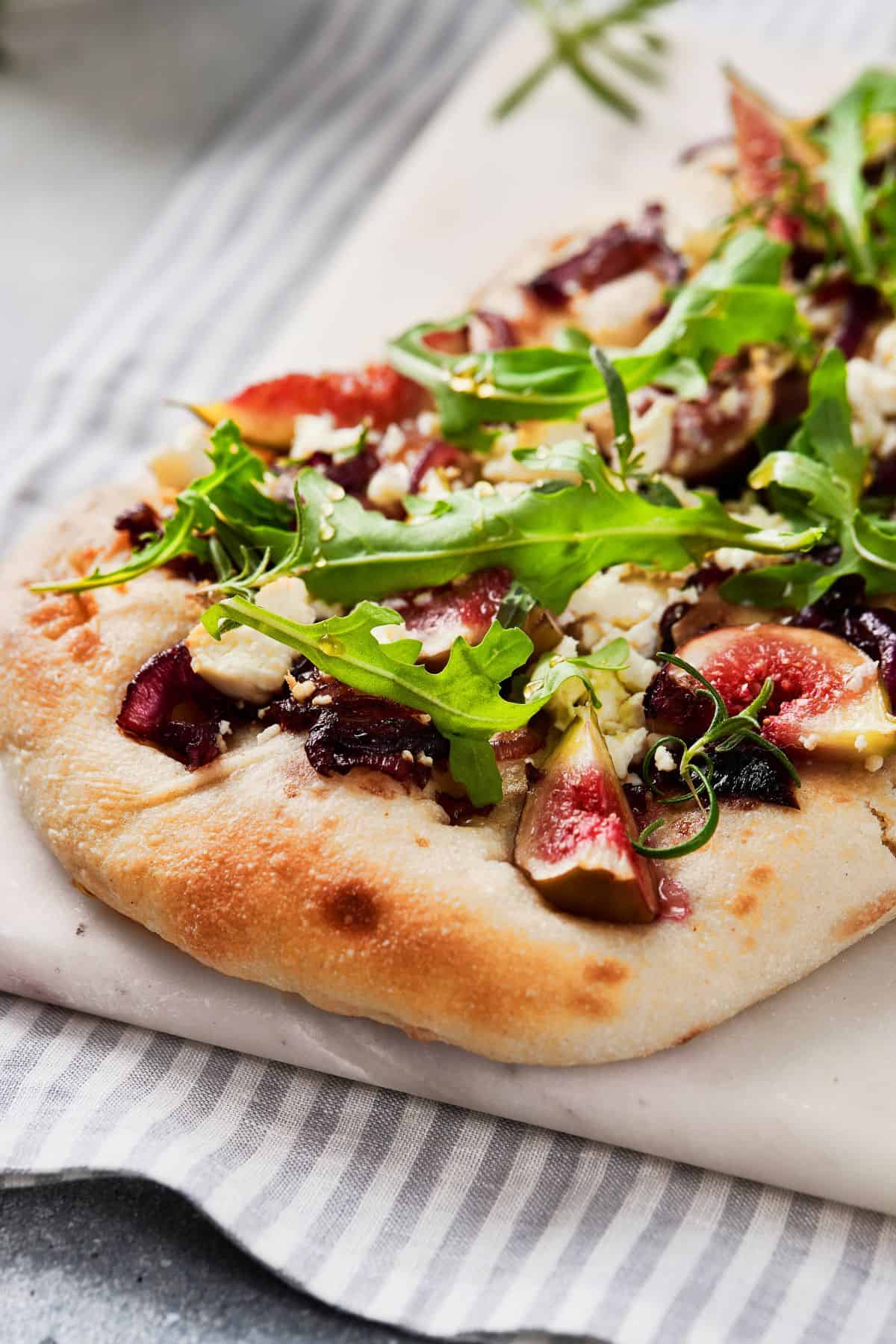 Close-up photo of flatbread garnished with arugula and goat cheese, surrounded with ingredients on a tablecloth underneath.