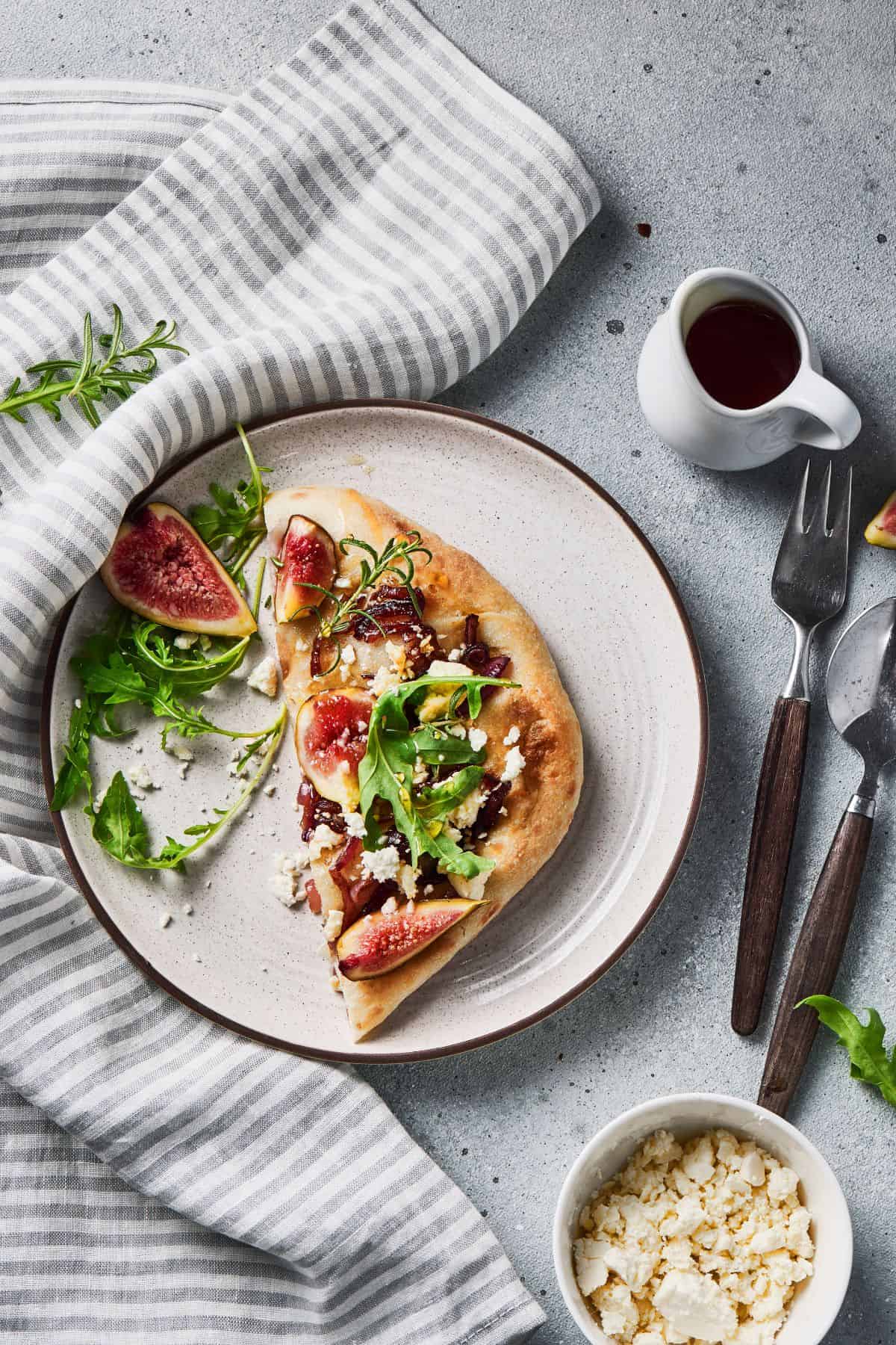 Sliced flatbread on a plate with a fork on the side, garnished with figs, caramelized onions, goat cheese, and arugula.