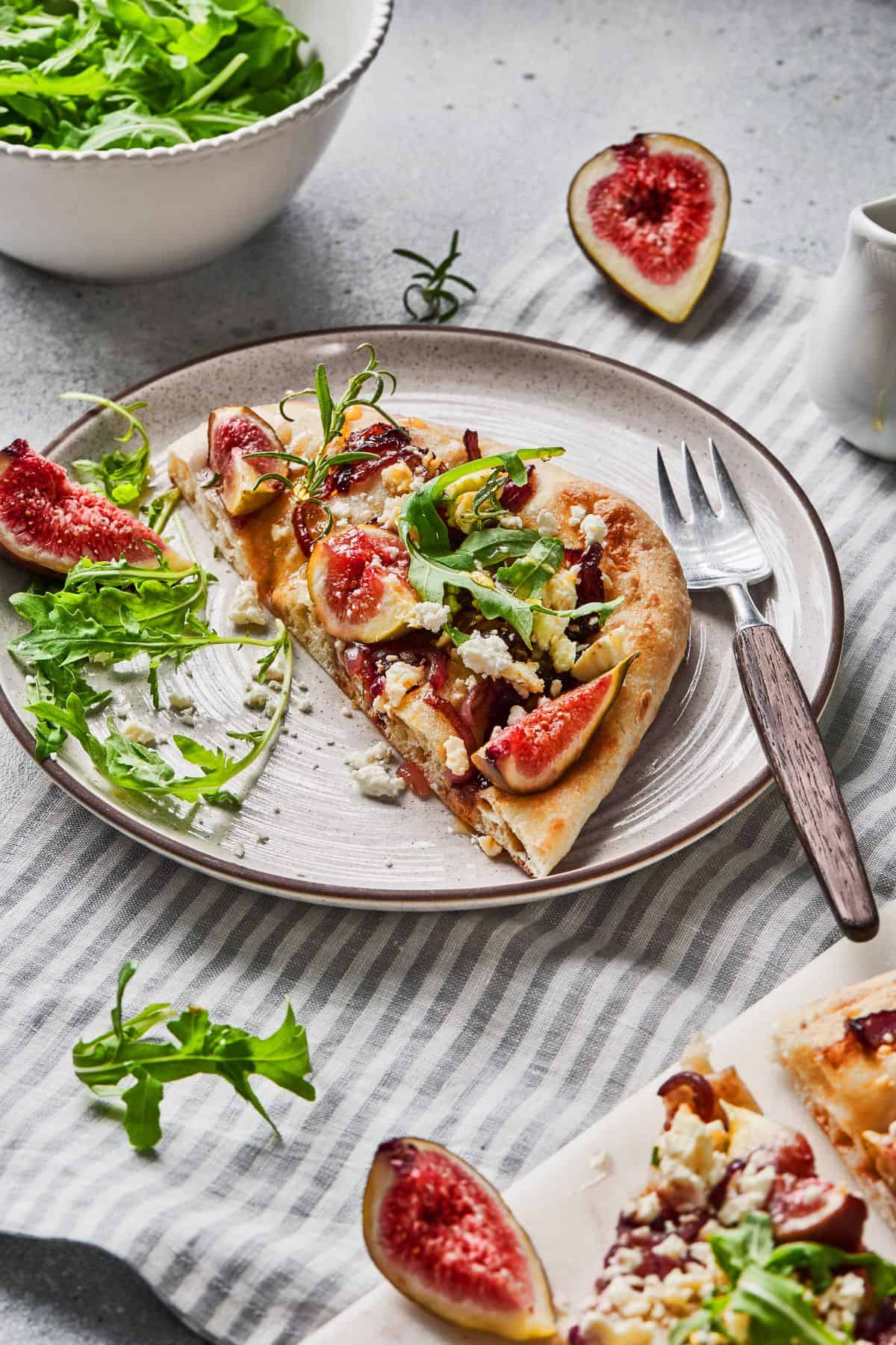 Sliced flatbread on a plate with a fork on the side, garnished with figs, caramelized onions, goat cheese, and arugula.