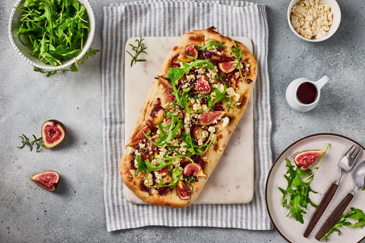 Served flatbread garnished with arugula and goat cheese, surrounded with ingredients on a tablecloth underneath.